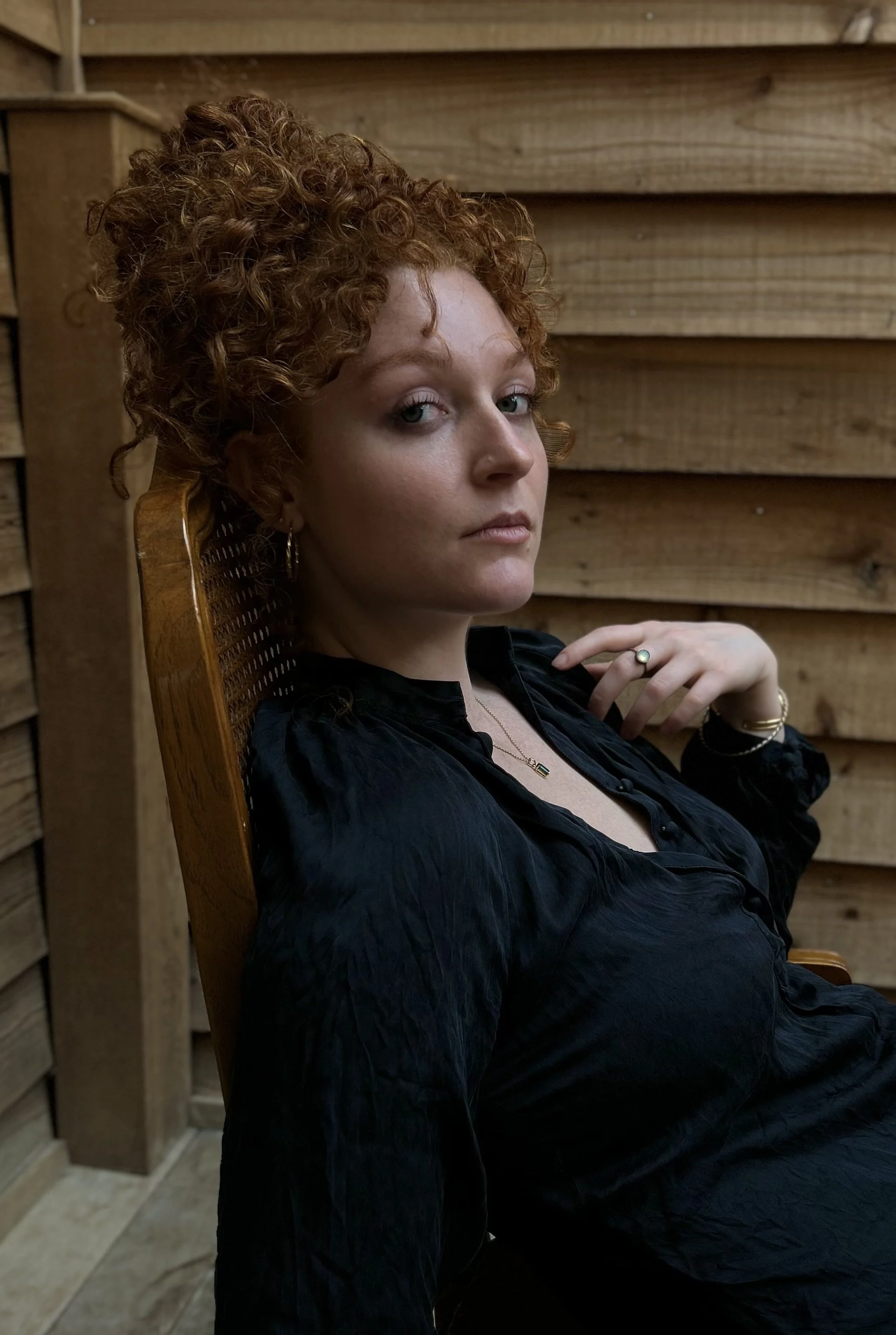 A woman with curly red hair sitting on a wooden chair, wearing a black shirt and jewelry, against a wooden wall background.