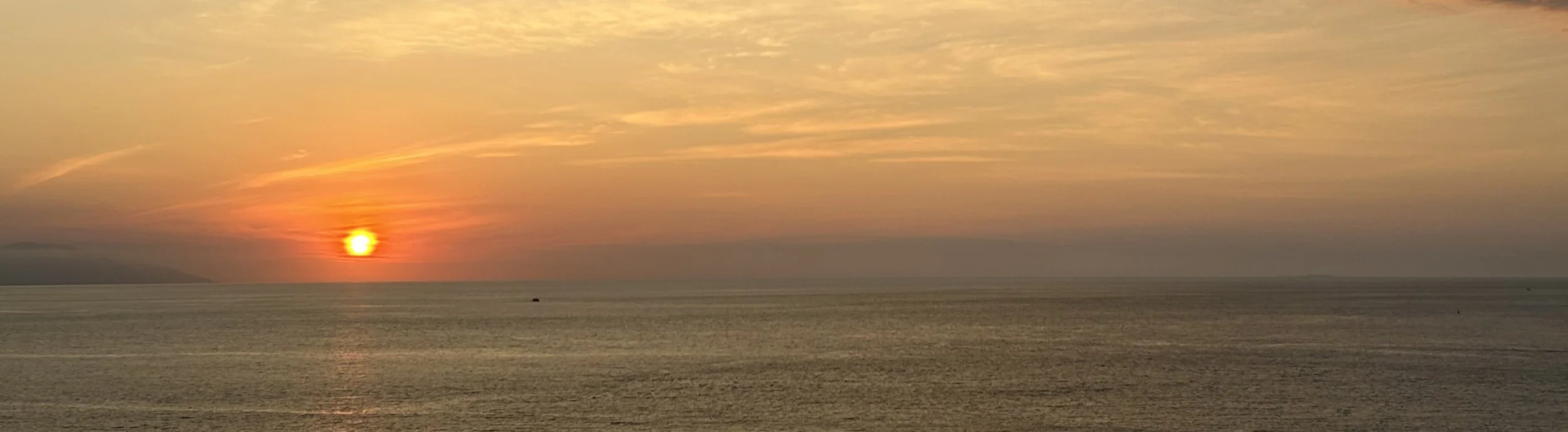Sunset over the ocean with a cloudy sky, distant land on the horizon, and a small boat on the water.