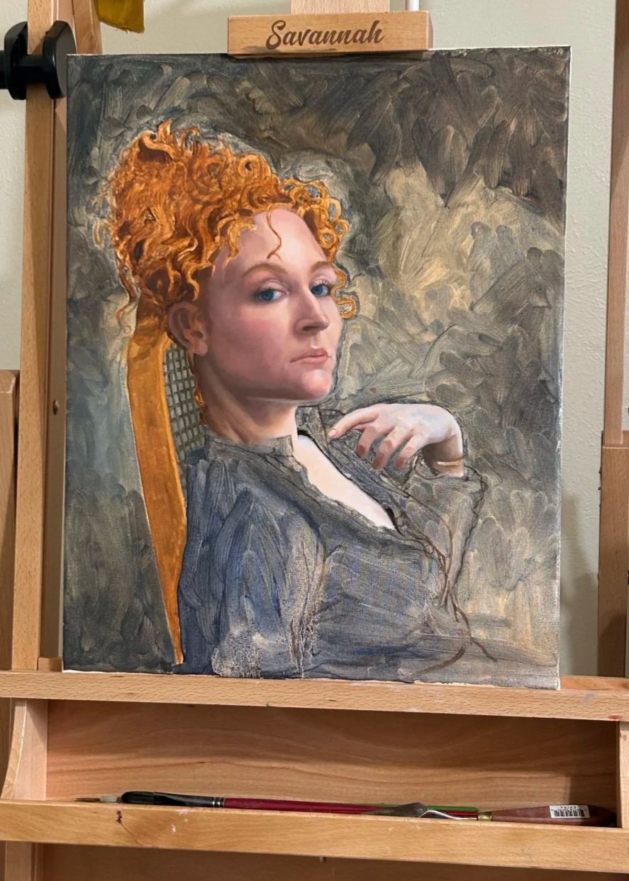 A painting of a woman with curly red hair, blue eyes, and fair skin, looking over her shoulder with her hand on her chest. She wears a dark top, and the background is a textured, earthy tone. The canvas is a work in progress sitting upon an easel.