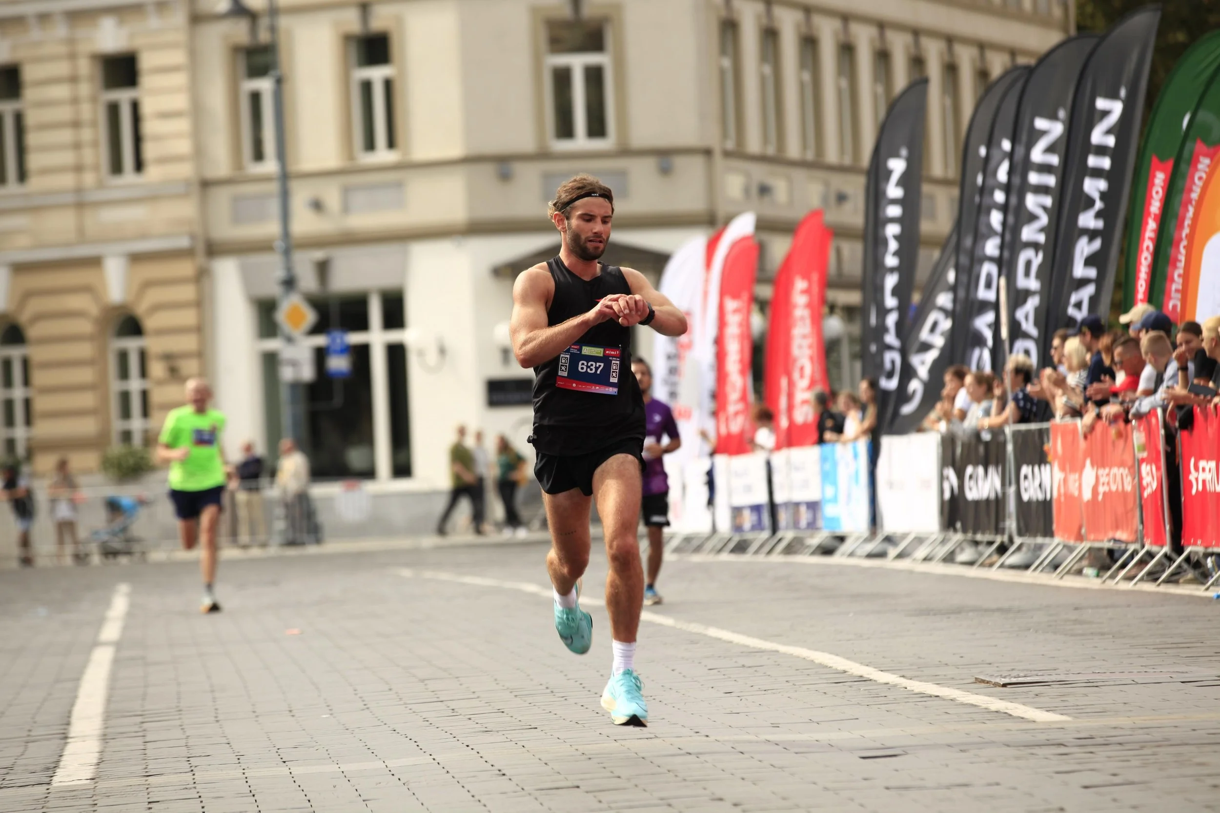 A man running in a race, wearing a black tank top and shorts, with a race number 637 attached to his shirt. He is looking at his wristwatch while running on a paved city street, with spectators behind barriers and colorful flags on the side. The background features buildings and other runners.