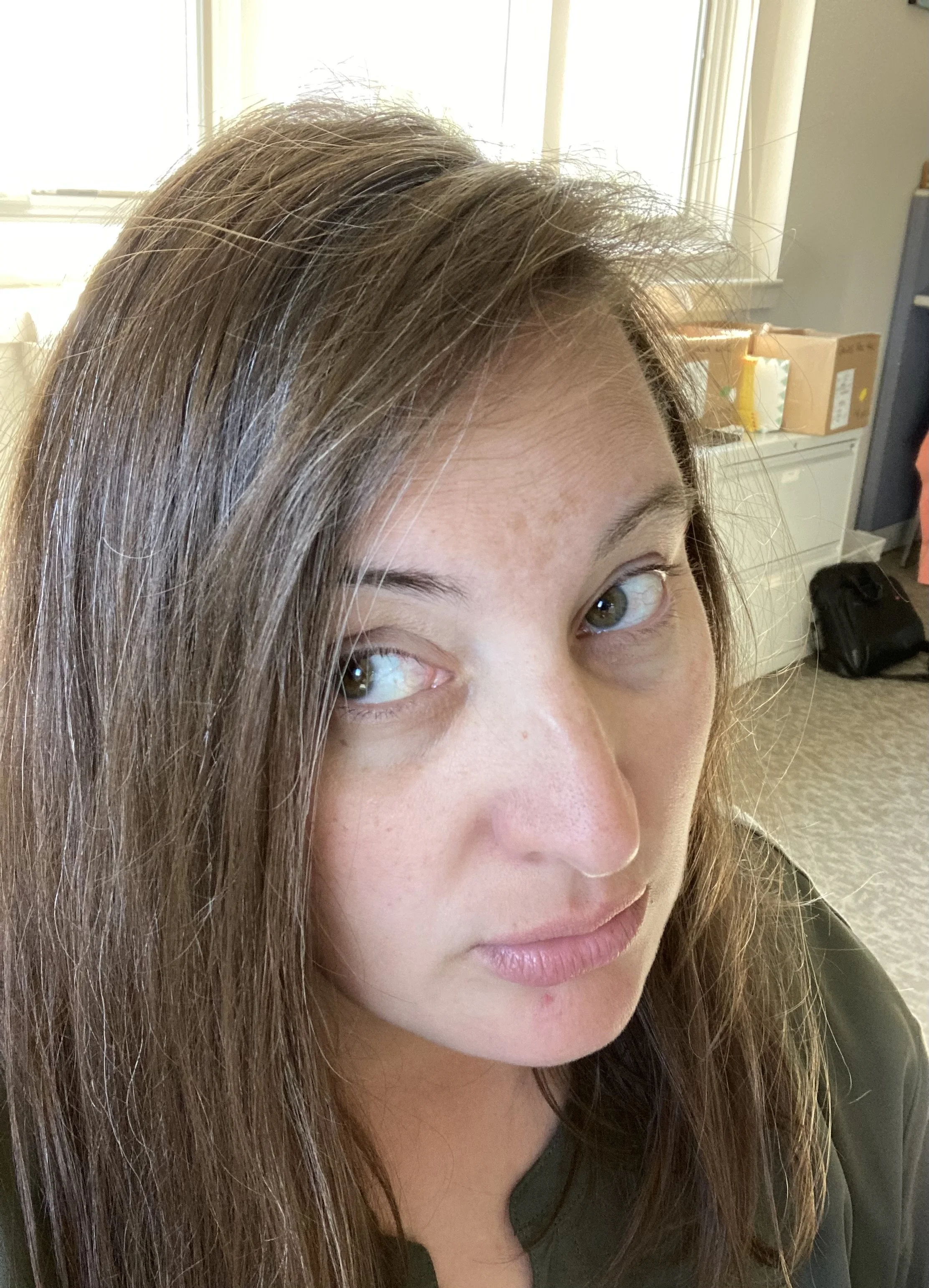 Close-up of a woman's face with long brown hair, green eyes, and light skin in an indoor setting near a window with boxes in the background.