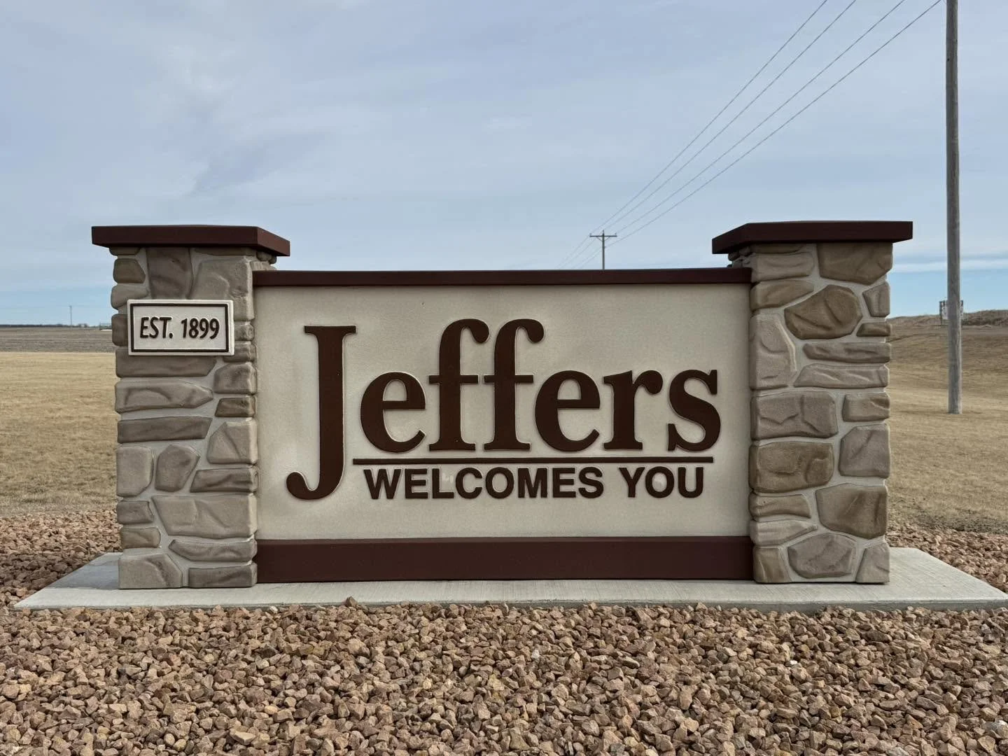 A stone and stucco welcome sign for Jeffers, established in 1899, with a brown border and text that reads "Jeffers Welcomes You." The sign is on a gravel lot with a rural background and utility poles in the distance.