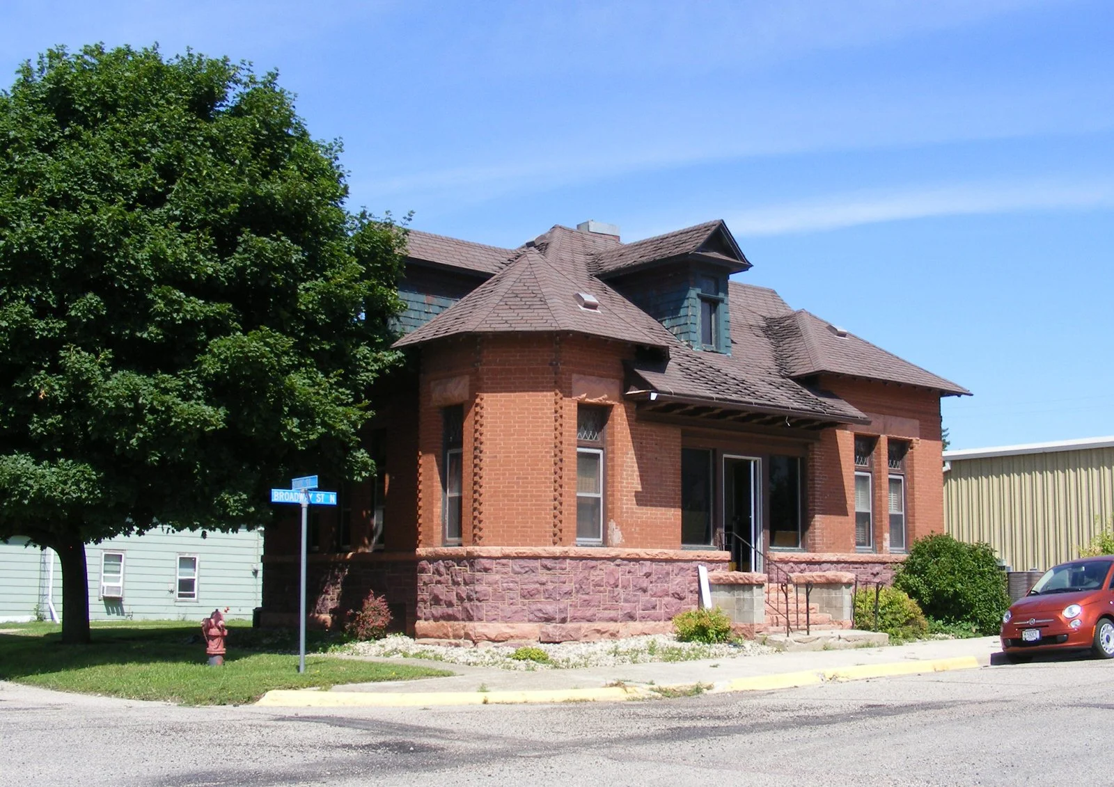 A brick house with a turret on a city street, a large tree, a street sign reading Broadway St N, a red fire hydrant, and a red car parked nearby.