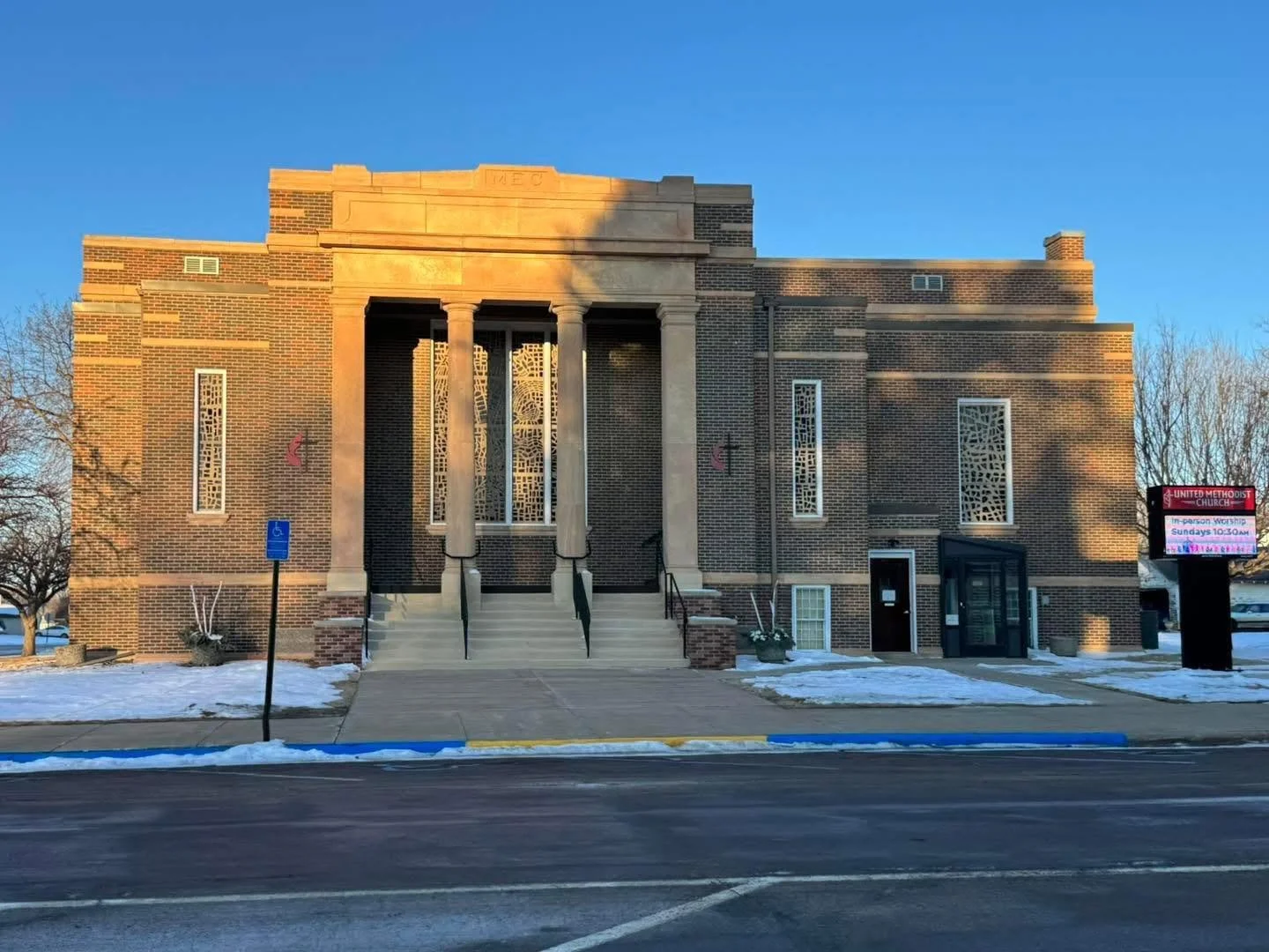 Front view of a brick church building with four tall columns at the entrance, glass doors, decorative windows, snow on the ground, and a digital signboard on the right side displaying church information.