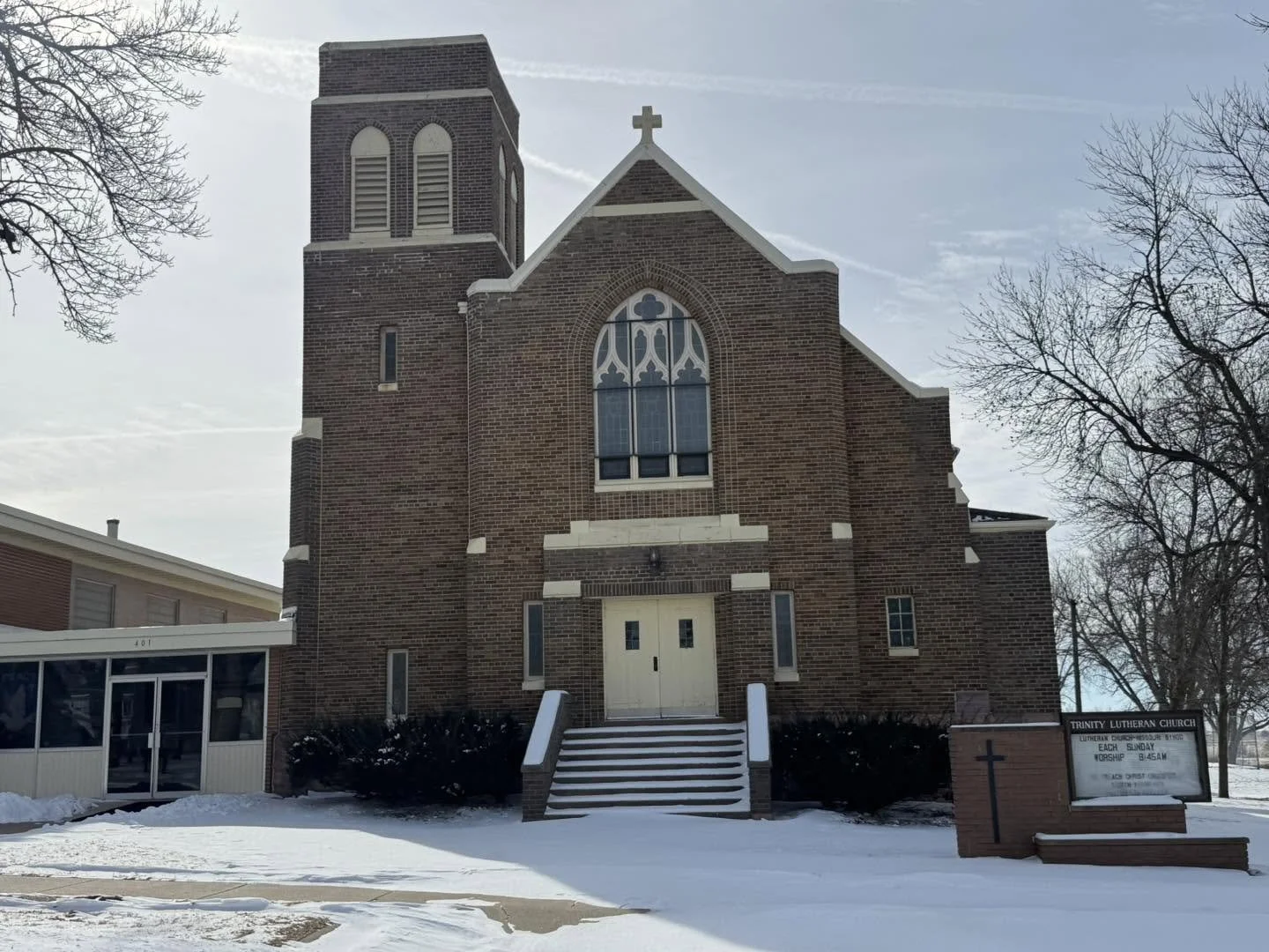 A brick church with a tall steeple, large stained glass window, and steps leading to a white door, surrounded by leafless trees and snow on the ground.