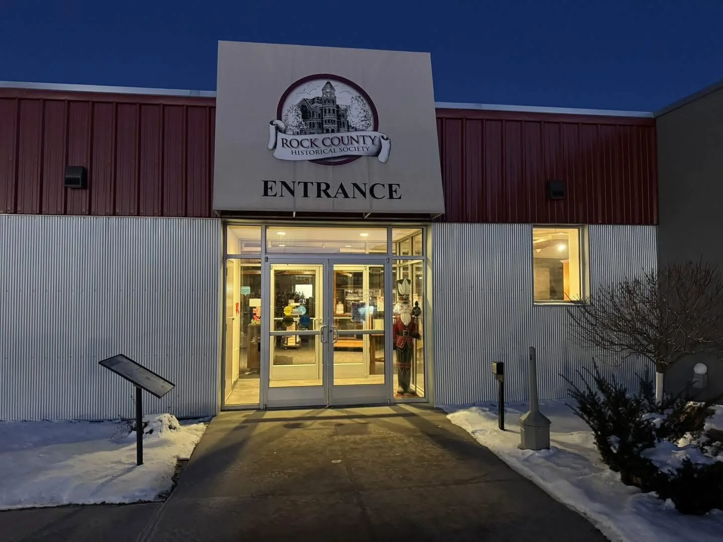 The entrance of the Rock County Historical Society building with a sign above the door reading 'ENTRANCE'. The building has brown and gray exterior walls, glass doors, and illuminated interior, with snow on the ground and a small bush on the right side.