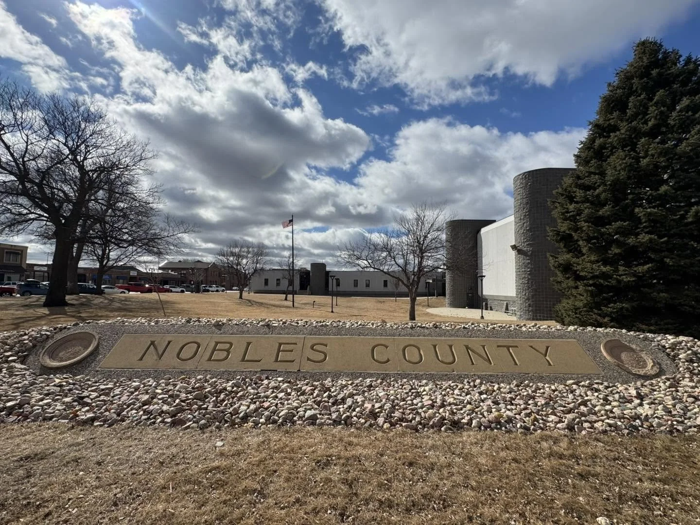 Stone sign reads 'Nobles County' in front of a park with trees, a grassy area, a flagpole with the American flag, and buildings in the background under a partly cloudy sky.
