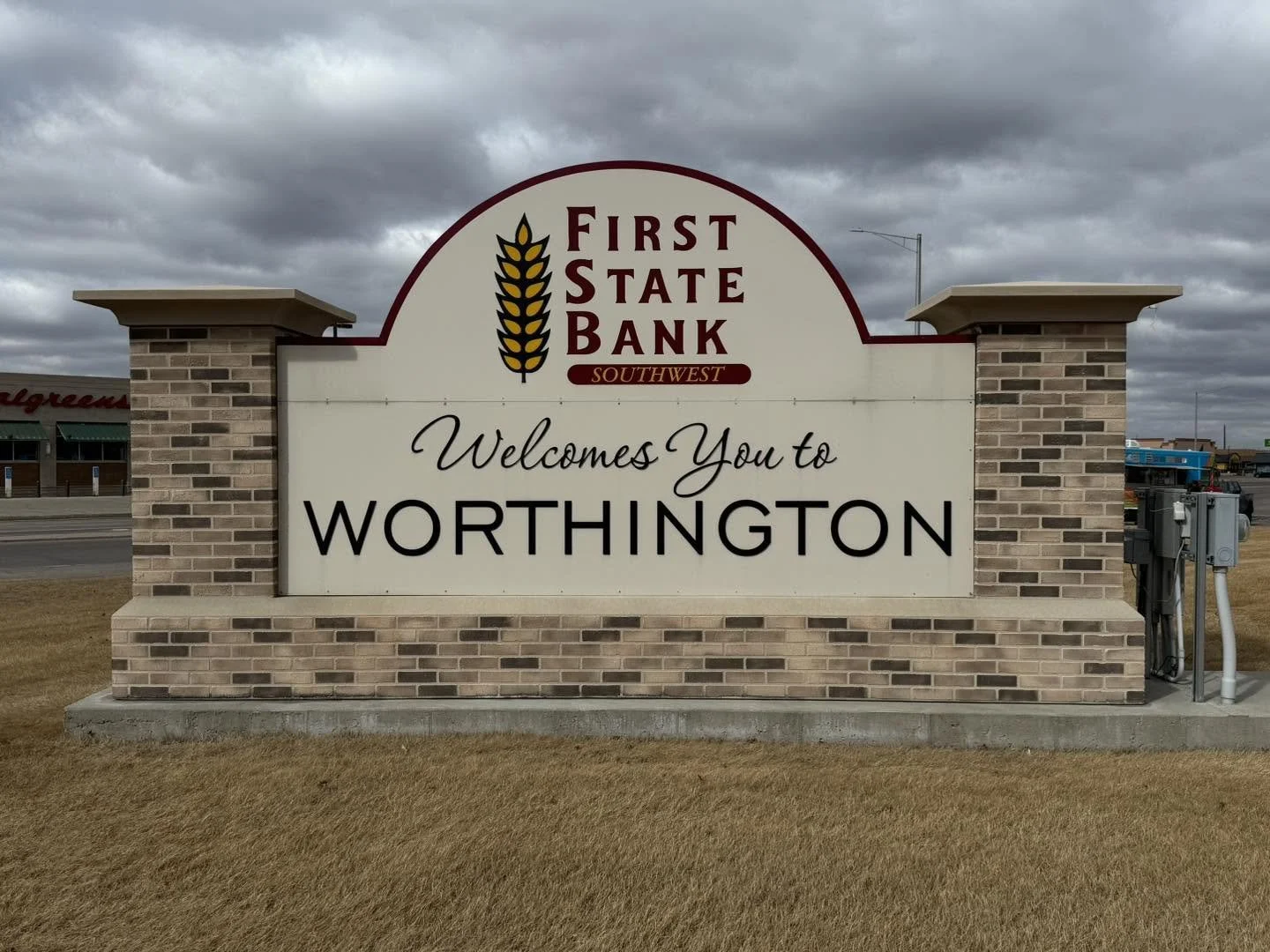 Welcome sign for Worthington, Ohio with the First State Bank Southwest logo and a wheat icon, on a brick and concrete base, on a grassy area under a cloudy sky.