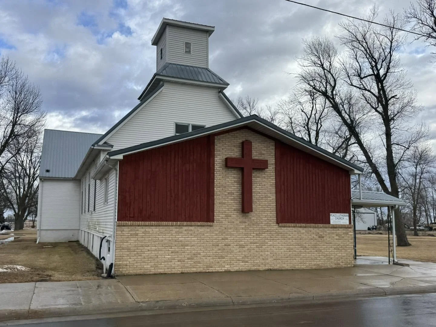Exterior of a church building with a large cross on the front. The church has white siding, a brick section, and red wooden panels. A small sign reads 'Peace Lutheran Church,' and the surroundings include leafless trees, a sidewalk, and an overcast sky.