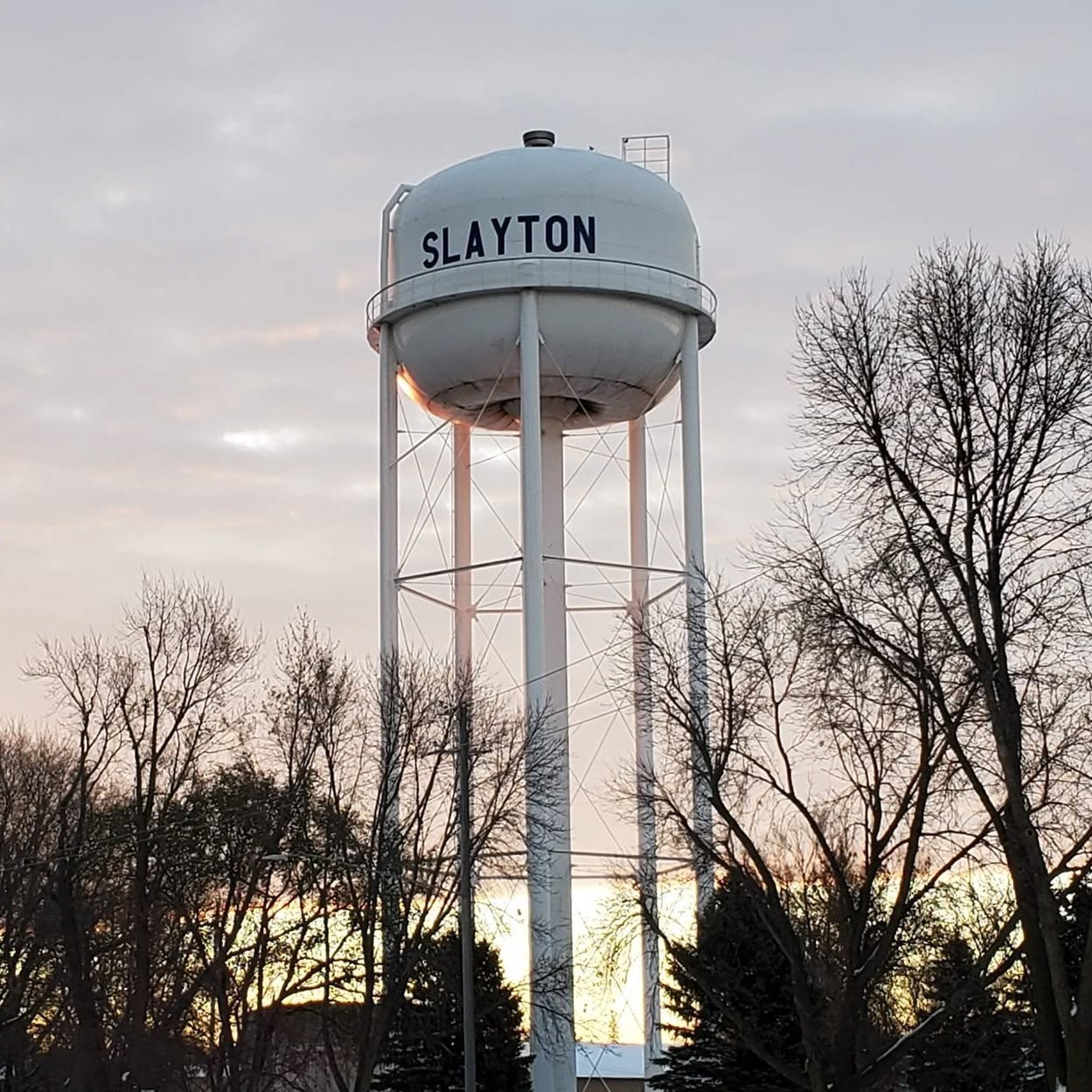 A tall white water tower with the word 'SLAYTON' on it, surrounded by leafless trees during sunset or sunrise.