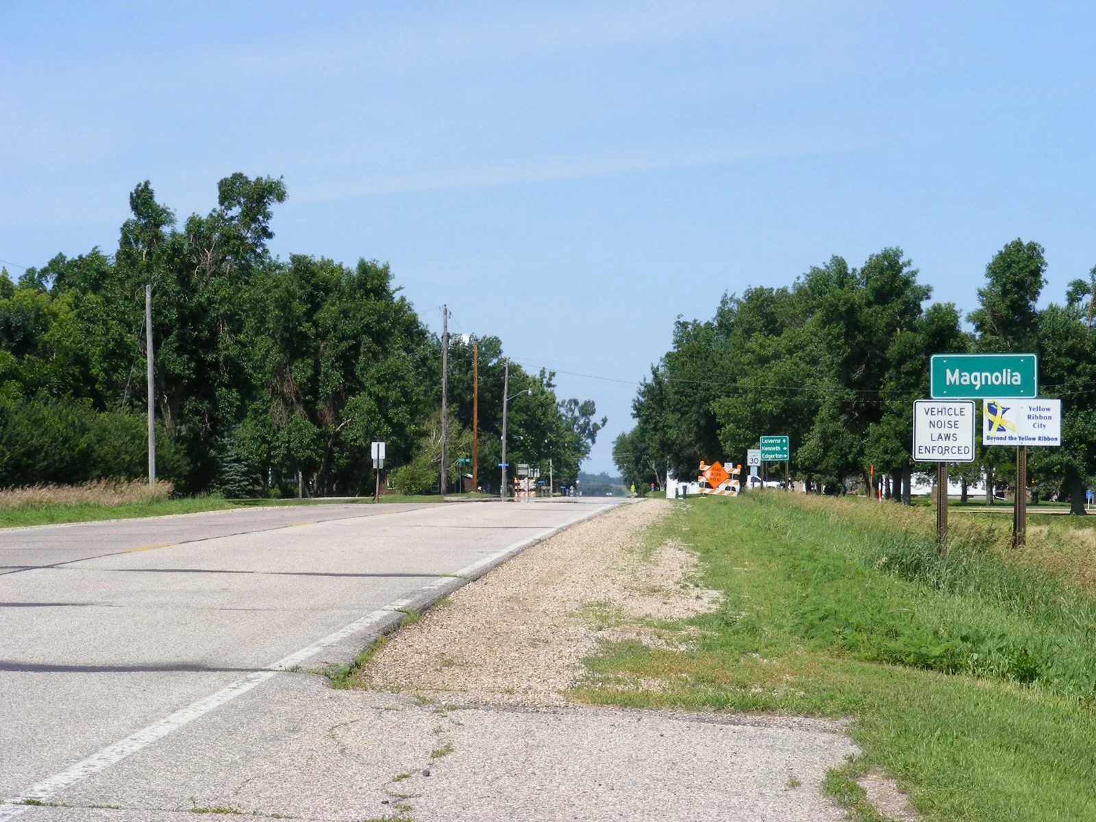 A rural street in Magnolia, with trees lining the road and traffic signs on the right, including a sign for vehicle noise laws enforcement and a Yellow Ribbon City sign.
