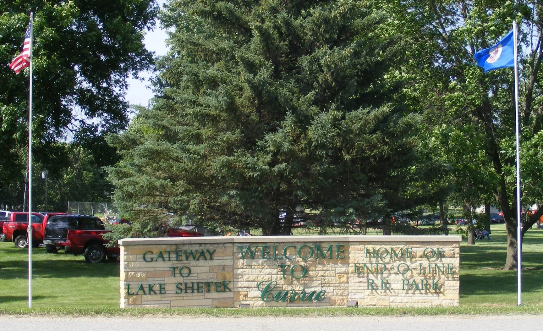 Stone sign that reads "Gateway to Lake Shetek, Welcome Home of End of Line R.R. Park" in front of a large evergreen tree, with flags of the United States and Minnesota on flagpoles, and parked cars visible in the background.
