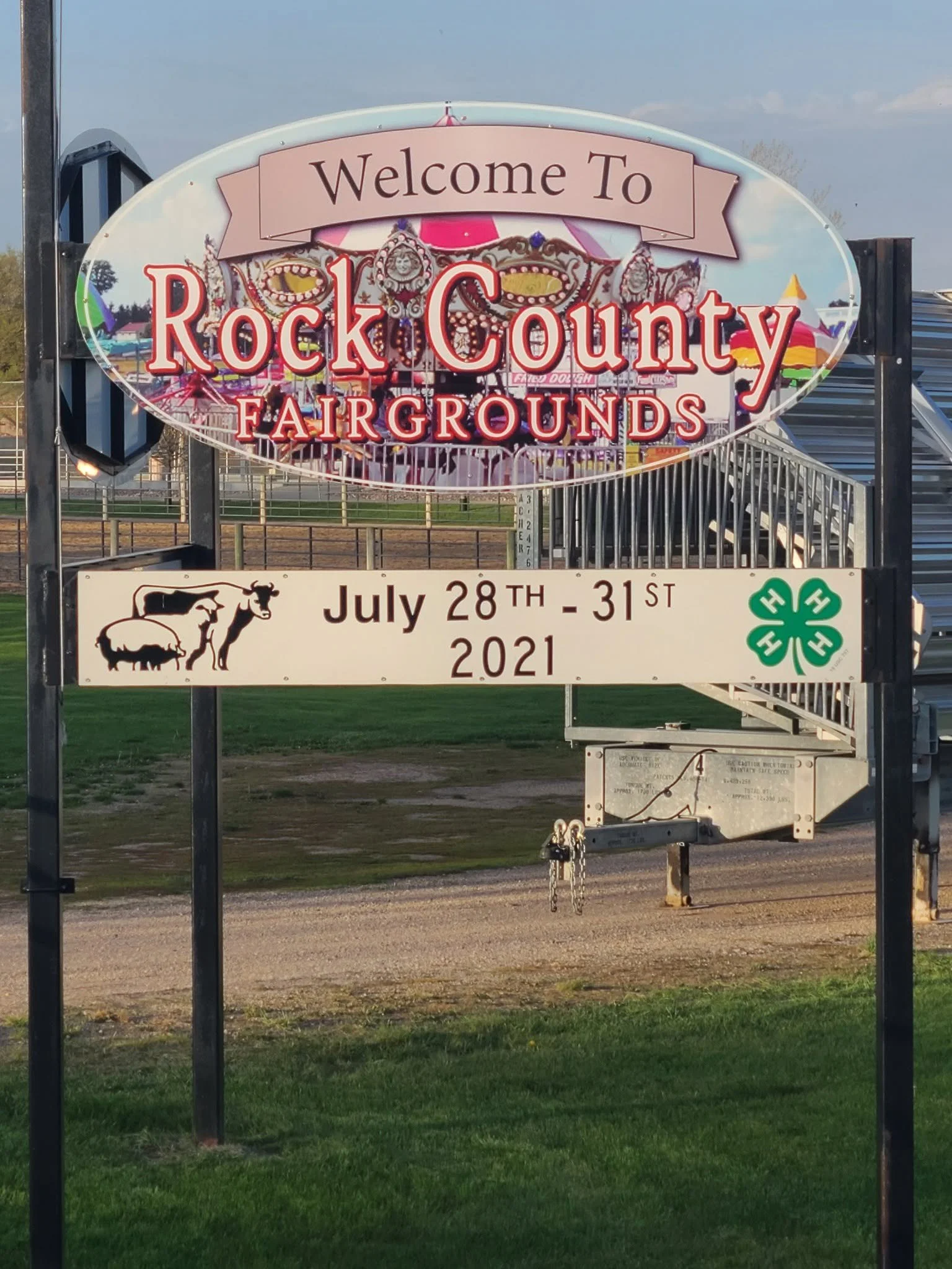 Sign welcoming visitors to Rock County Fairgrounds, with amusement park rides and carnival games in the background, dated July 28th-31st, 2021.