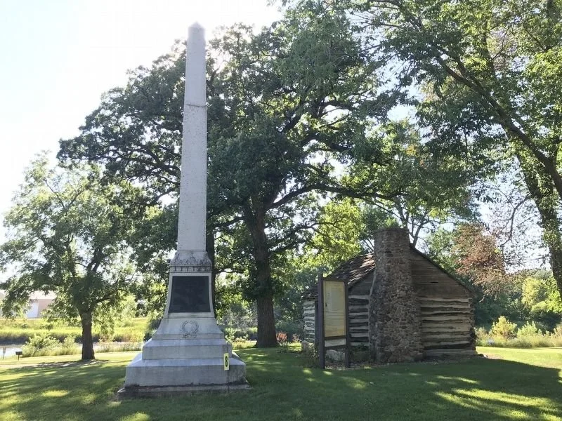 An outdoor scene featuring a white obelisk monument and a small log cabin with a stone chimney, surrounded by trees and grassy area.