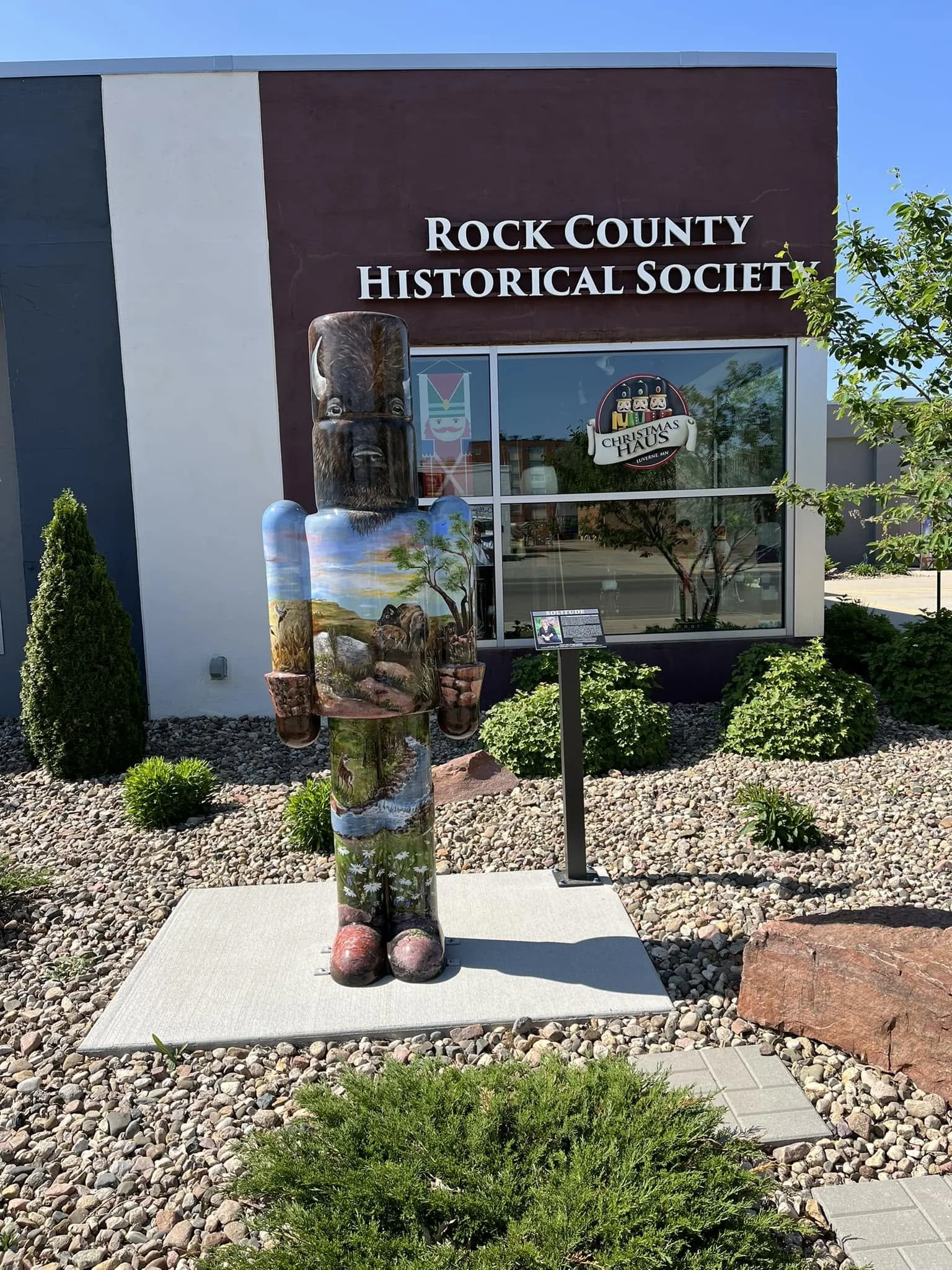 Statue in front of the Rock County Historical Society building, depicting a figure with a painted landscape design on the body and a carved head, standing on a concrete platform in a landscaped area.