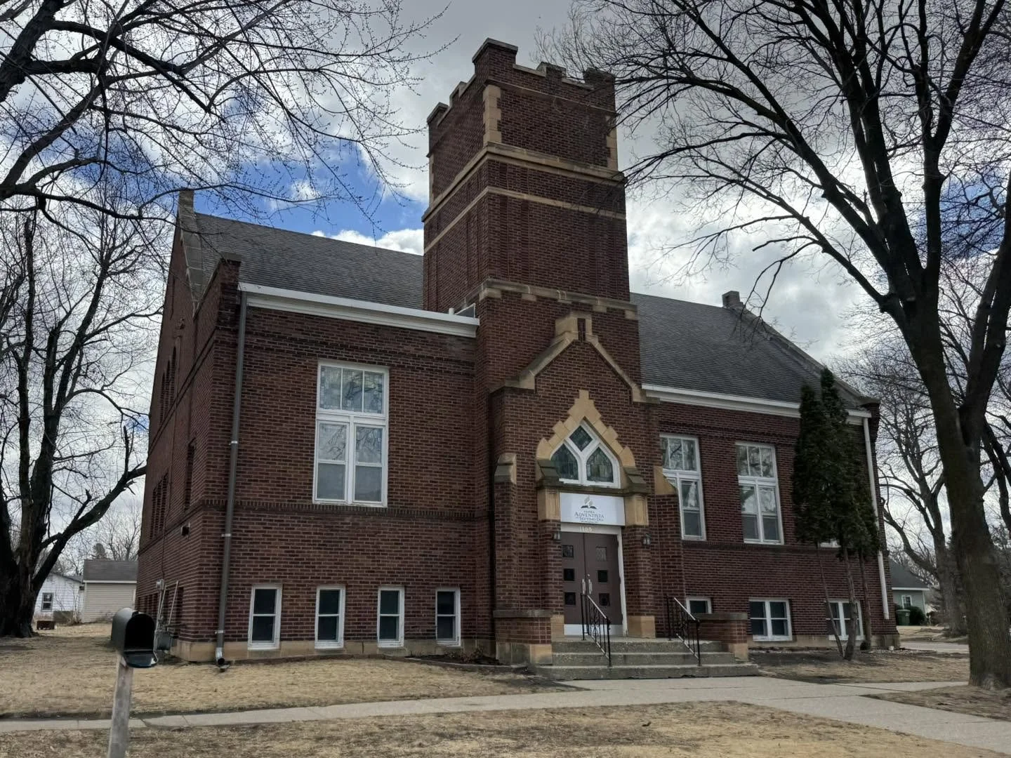 A red brick church with a tall steeple, pointed arch windows, and a set of steps leading to the entrance, surrounded by leafless trees on a cloudy day.