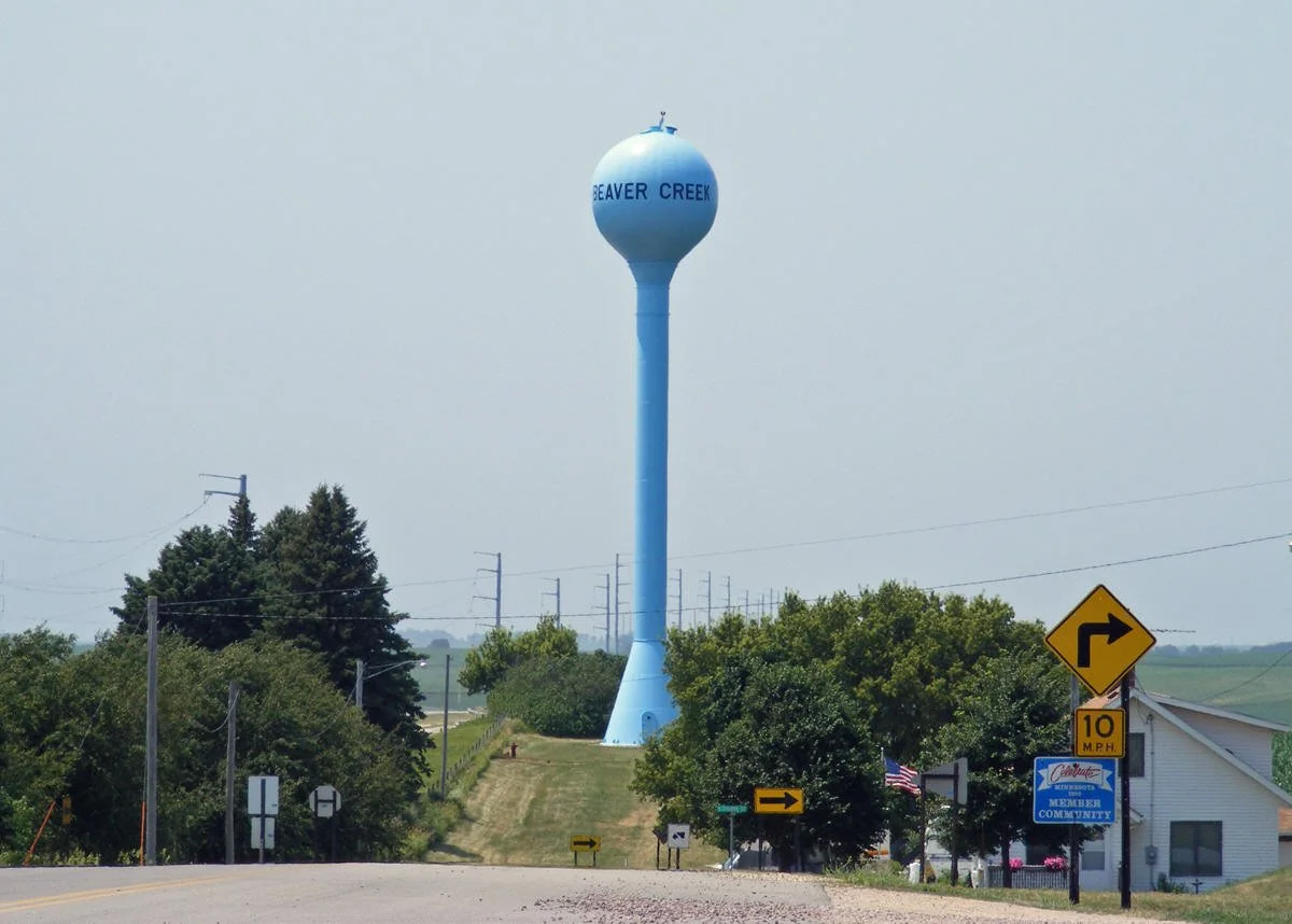 Water tower with the words 'Beaver Creek' on it, near a small road with directional signs, greenery, and a building with an American flag.