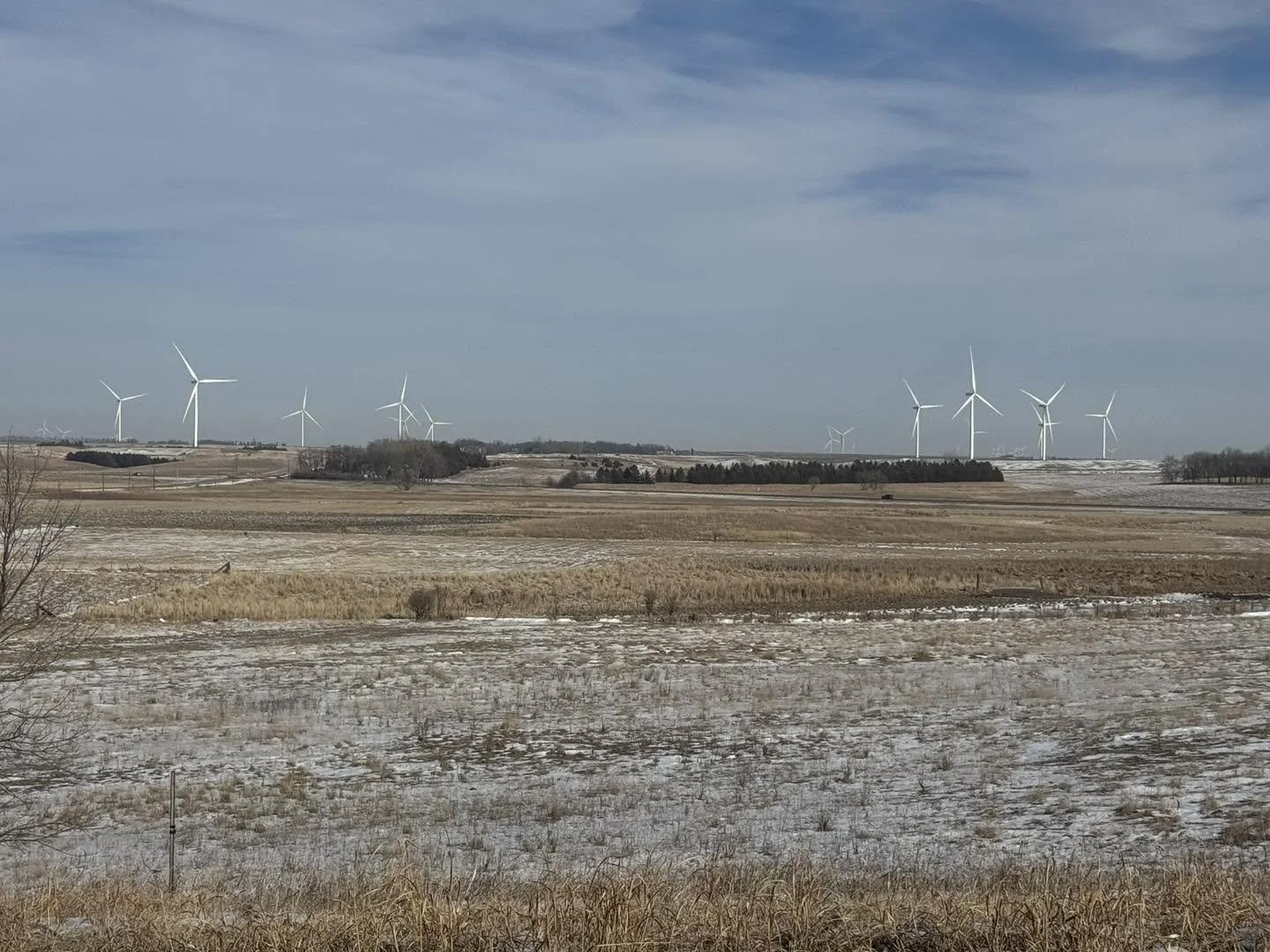 Open countryside with snow-dusted fields and a distant wind farm with numerous wind turbines on the horizon under a partly cloudy sky.