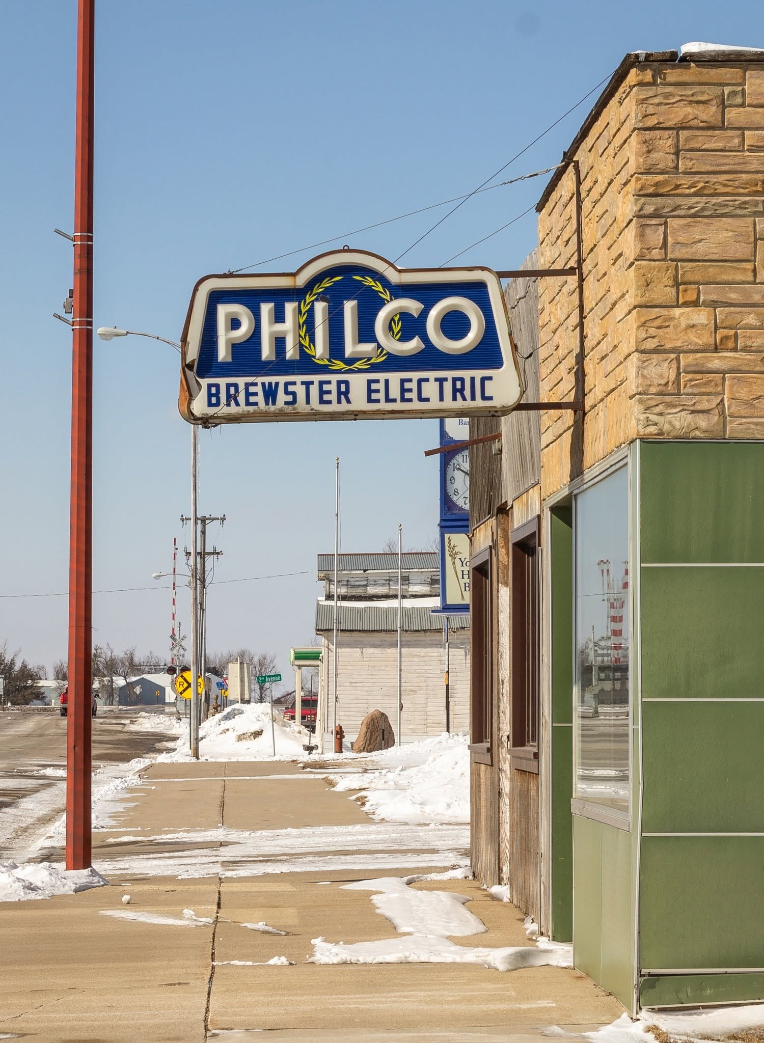 Street scene with snow on the sidewalk, a sign for Philo Brewster Electric, and a brick building with a green storefront.