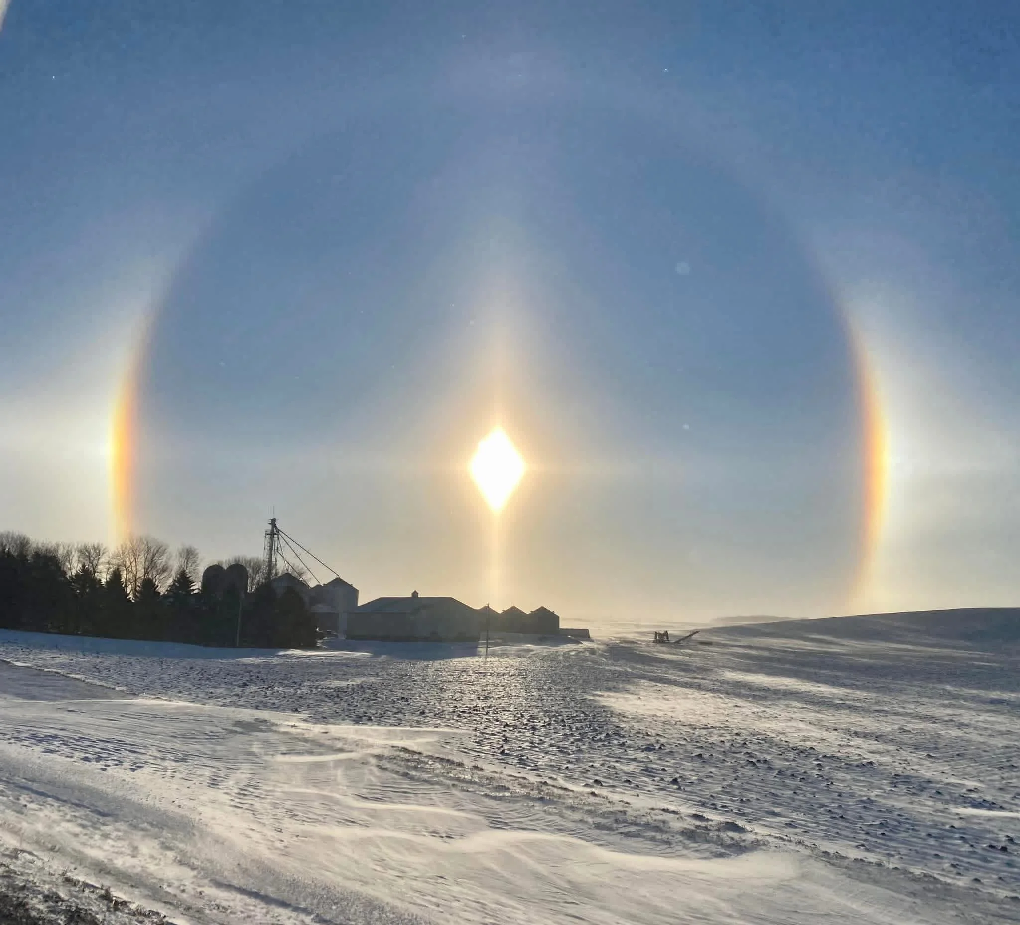 Sundog phenomenon in a winter landscape with snow-covered ground, trees, and houses, during sunset or sunrise.