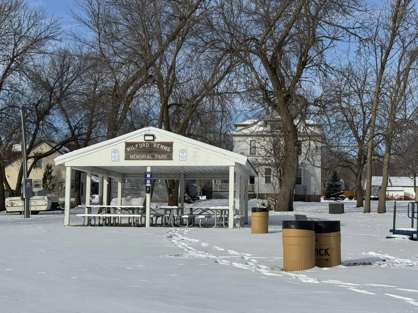 Snow-covered park with a pavilion that reads 'Milford Remme Memorial Park,' surrounded by leafless trees, trash bins, benches, and residential houses in the background.