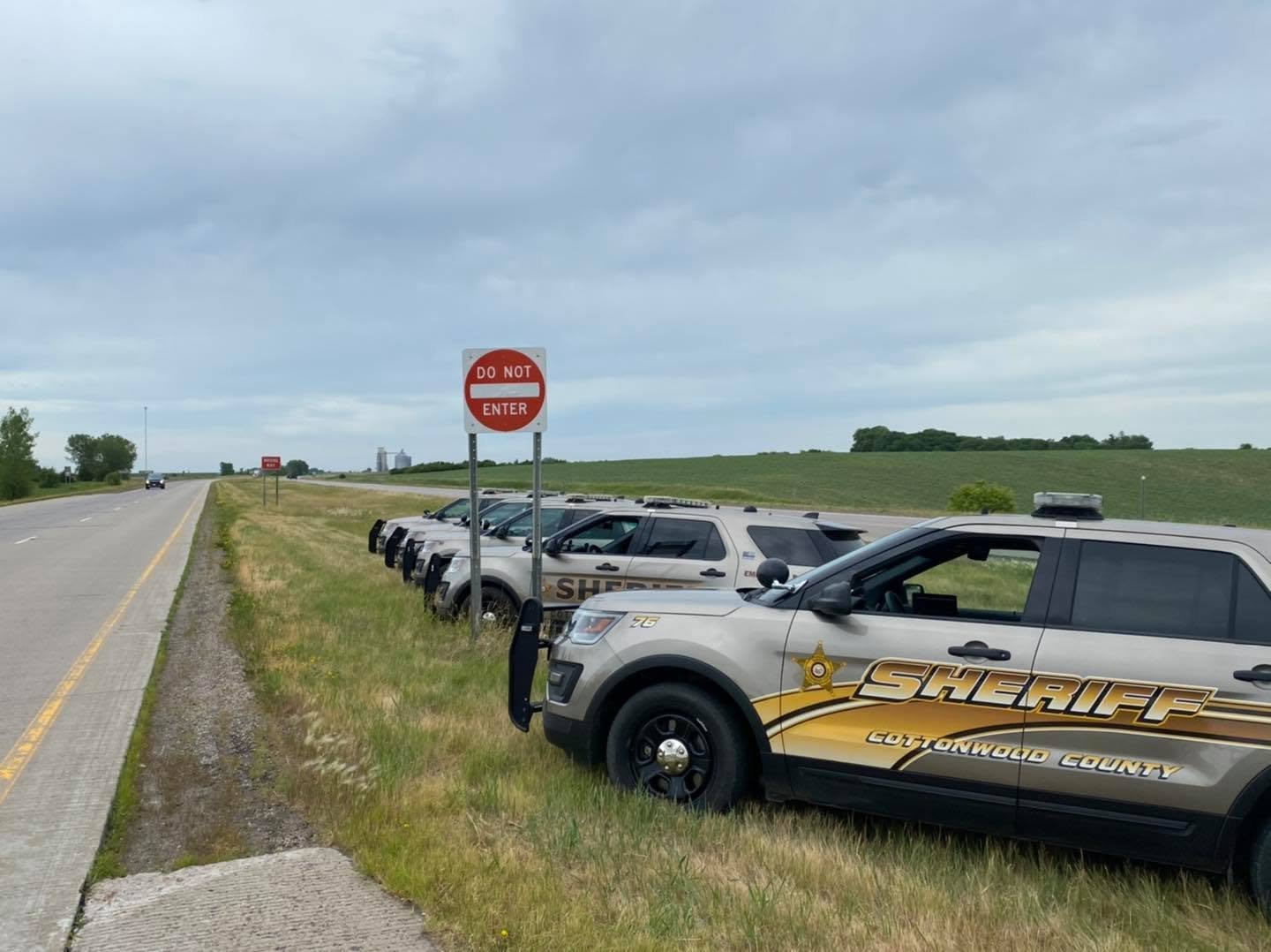 Multiple sheriff vehicles parked on the grassy shoulder of a highway next to a 'Do Not Enter' sign under a cloudy sky.