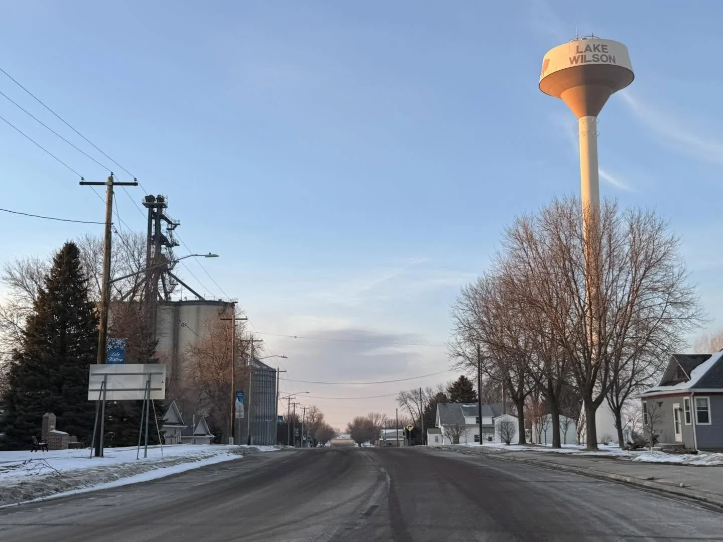 Empty street in a small town with snow on the ground, bare trees, houses, utility poles, and a water tower labeled 'Lake Wilson' in the distance.