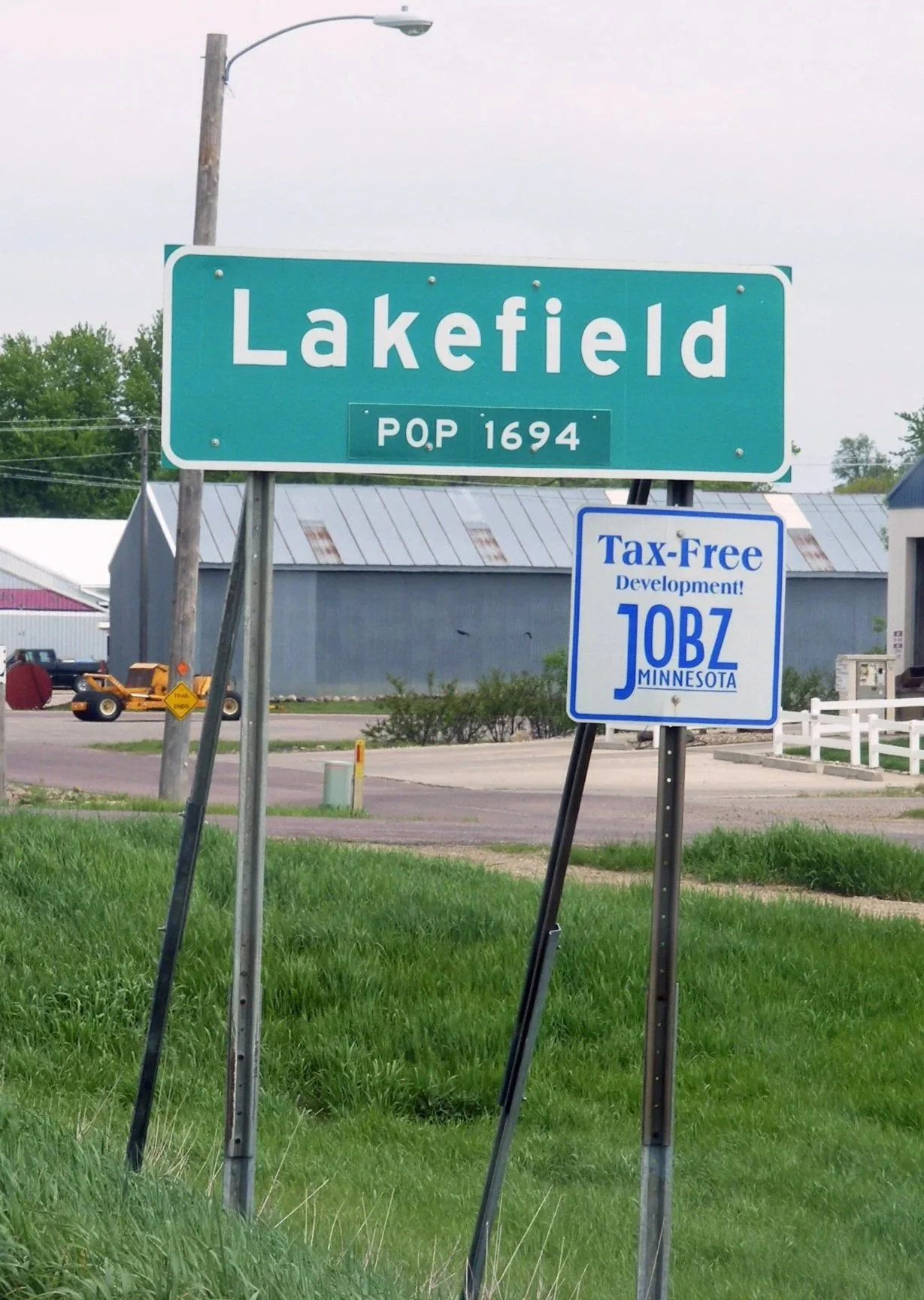 Green highway sign with white text reading 'Lakefield' and population 1694. Below it, a blue and white sign promotes tax-free development in Minnesota with the code J0BZ. The signs are on grass with some buildings and equipment in the background.