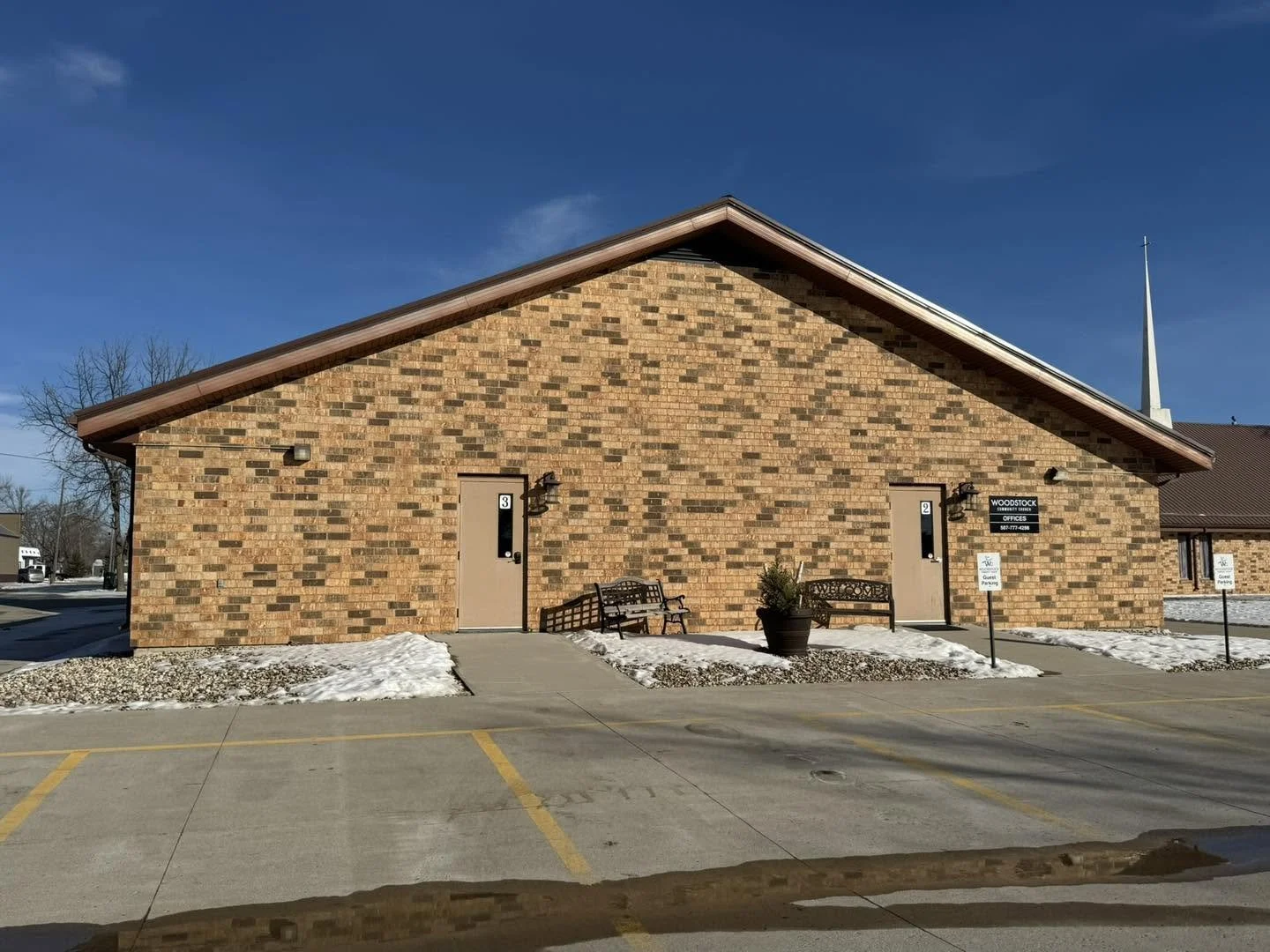 Brick building with two entrances labeled 2 and 3, benches, large flowerpot, snow on the ground, parking lot in front, signs indicating handicapped parking, clear blue sky, church steeple in background.