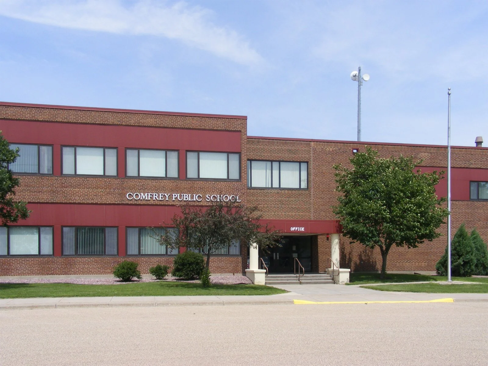 Front view of Comfrey Public School building with trees, flagpoles, and a sidewalk.