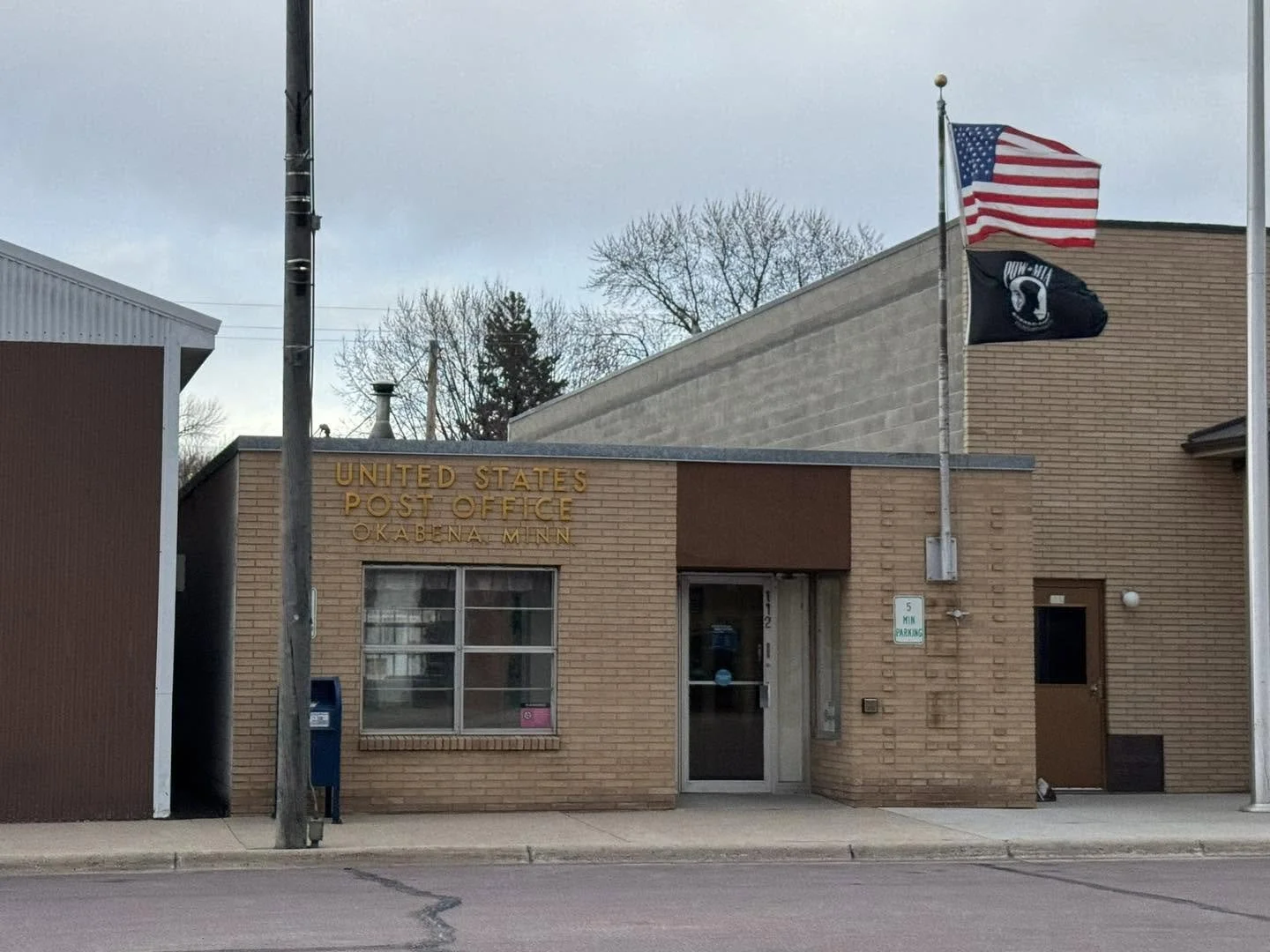 Front view of the United States Post Office building in Oaktina, Minnesota, with two flags (American and military), a sidewalk, and a parking sign.