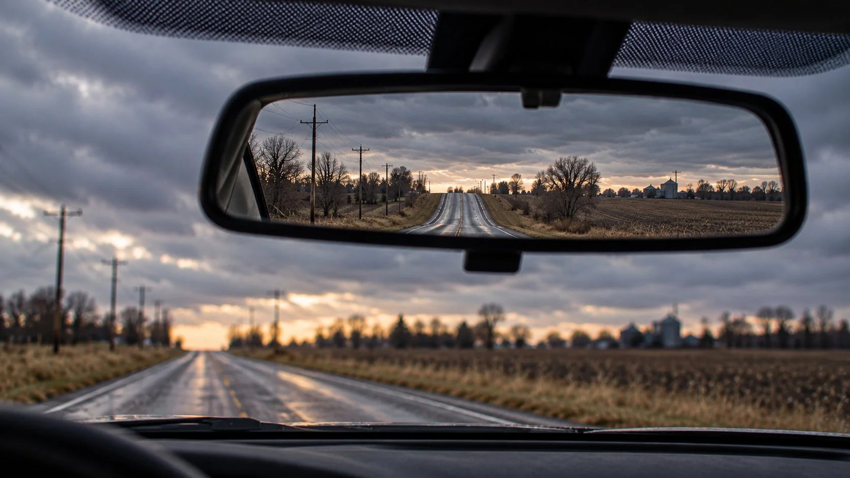 A view of an empty rural road seen through a car's rearview mirror, with a cloudy sky, power lines, and trees on both sides of the road.