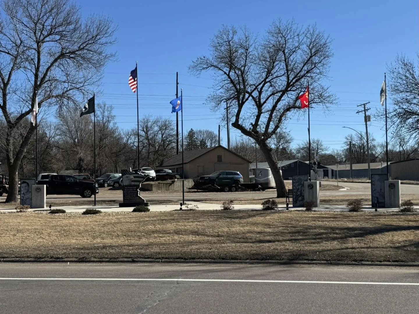 A memorial with seven flags, including the American flag, national flags, and flags on tall poles, in front of a small building, with a parking lot and several cars, trees without leaves, and utility poles in the background under a clear blue sky.
