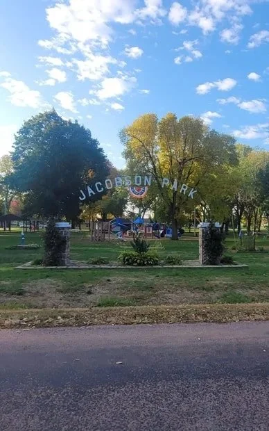 Entrance sign for Jacobson Park with trees, a pathway, and a playground in the background.