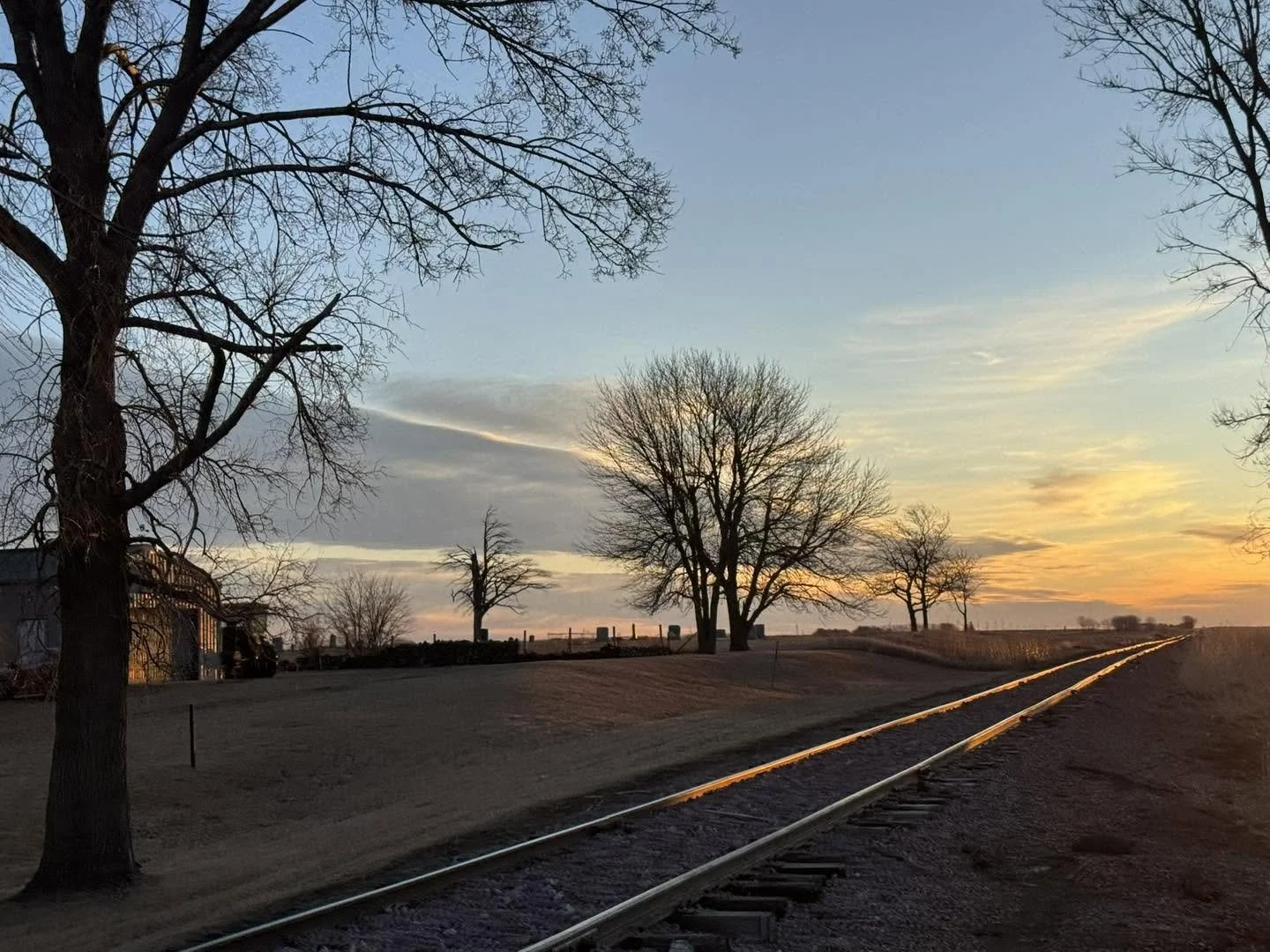 An empty train track running through a rural landscape at sunset with bare trees and a clear sky with some clouds.