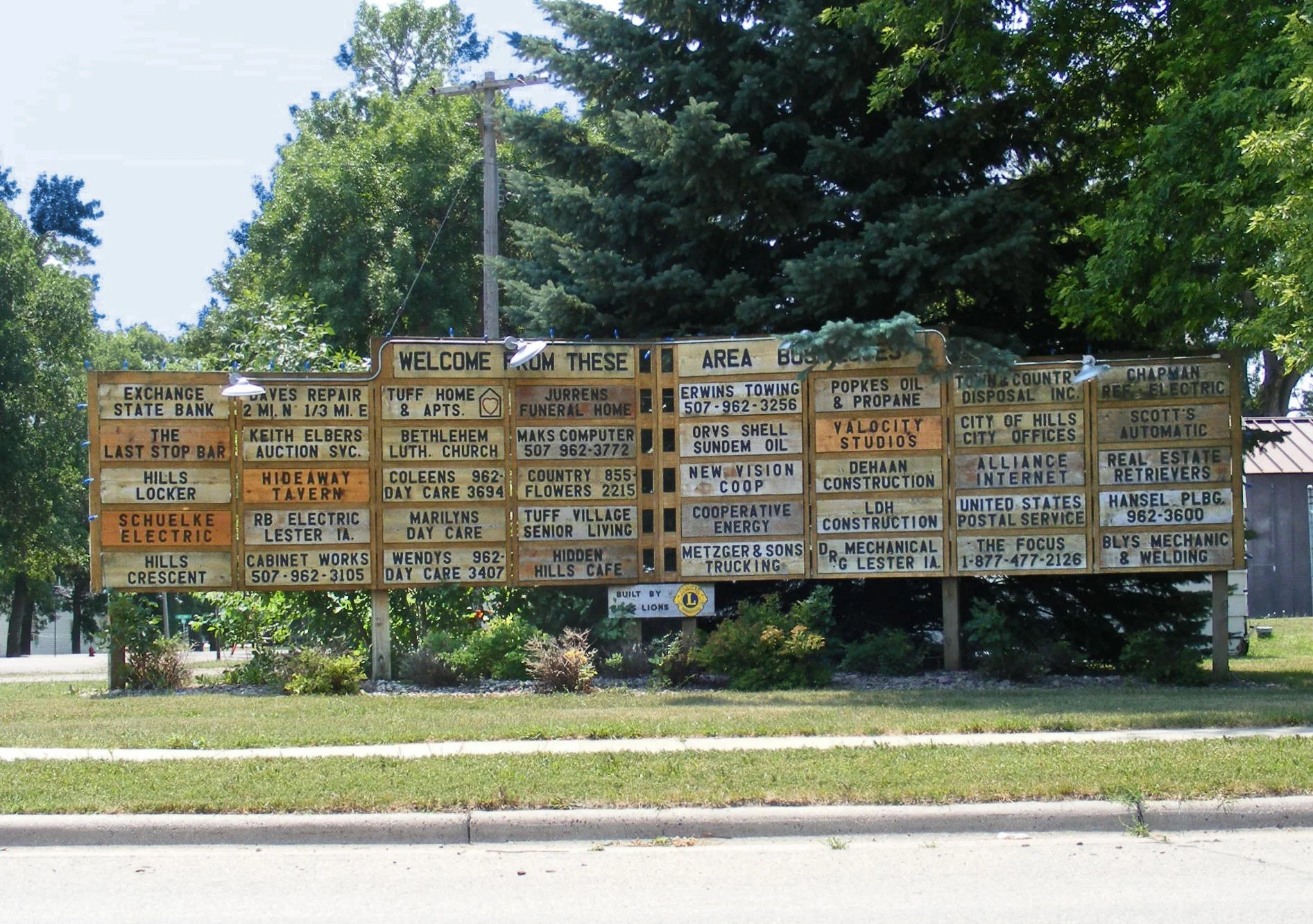 A large wooden billboard displays numerous small business advertisements with black text on a natural wood background, surrounded by green trees and a blue sky. The billboard includes messages such as "Welcome from these area businesses" at the top, and advertisements for various local companies like an electric service, a computer store, law office, and others.