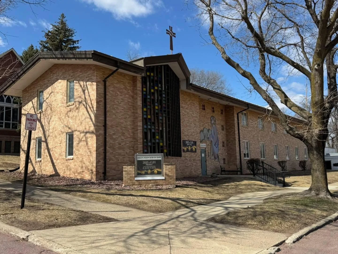 A brick church building with a cross on top, stained glass windows, and an exterior mural depicting Jesus with a lamb, alongside a sign for the Sheridan Jackson Quasquicentennial Center.