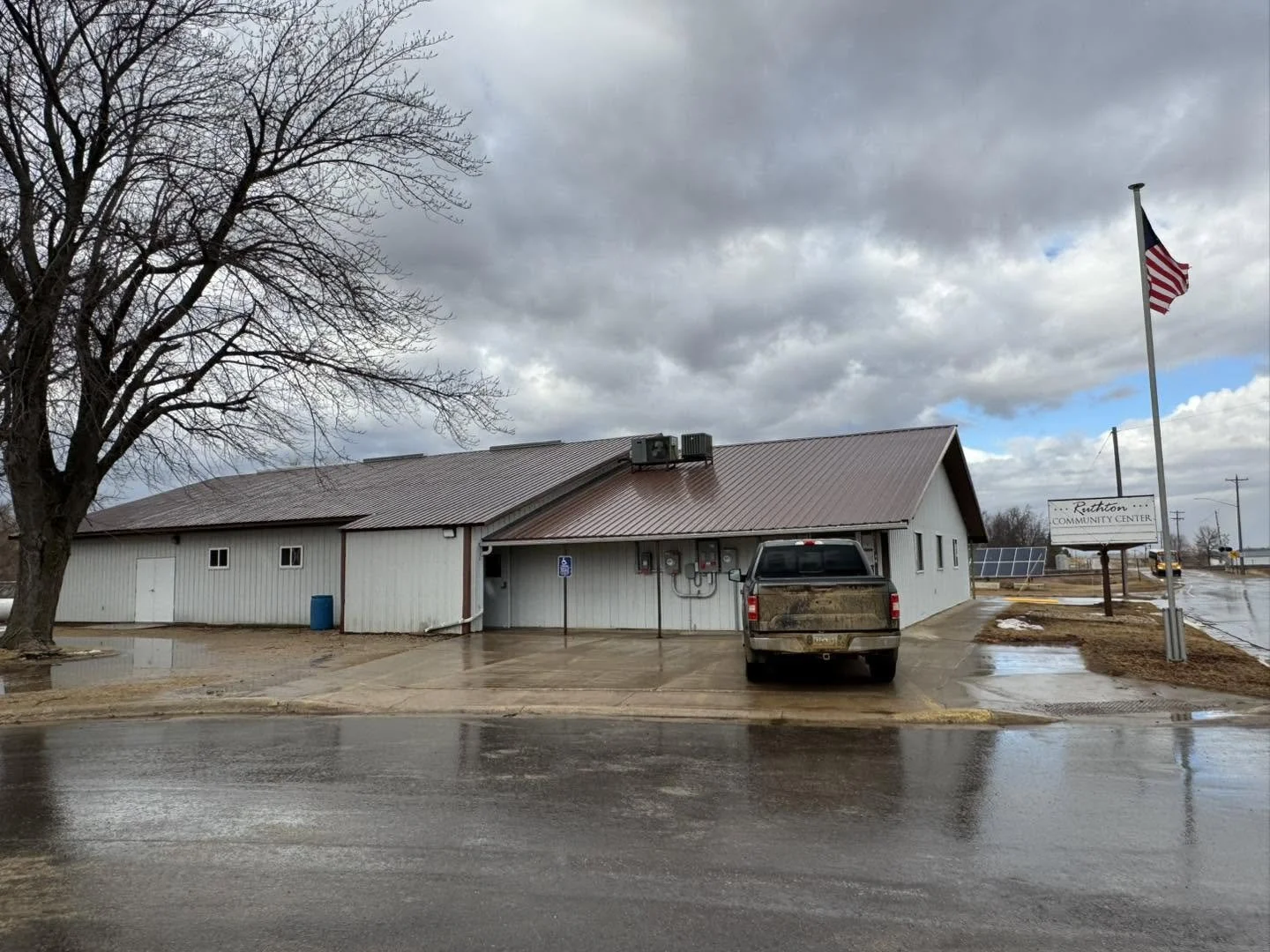 A building with a metal roof, parked truck, large tree without leaves, American flag, and a sign that reads 'Ralston Community Center,' on a rainy day.
