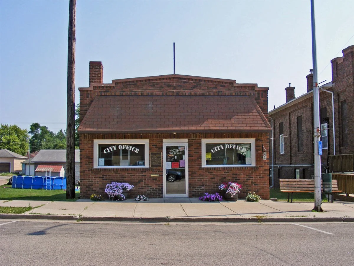 Brick city office building with two large front windows and a glass door. Flowers in pots are placed in front of the building. Sign on windows and door indicate it is a city office. A sidewalk and street are in the foreground.