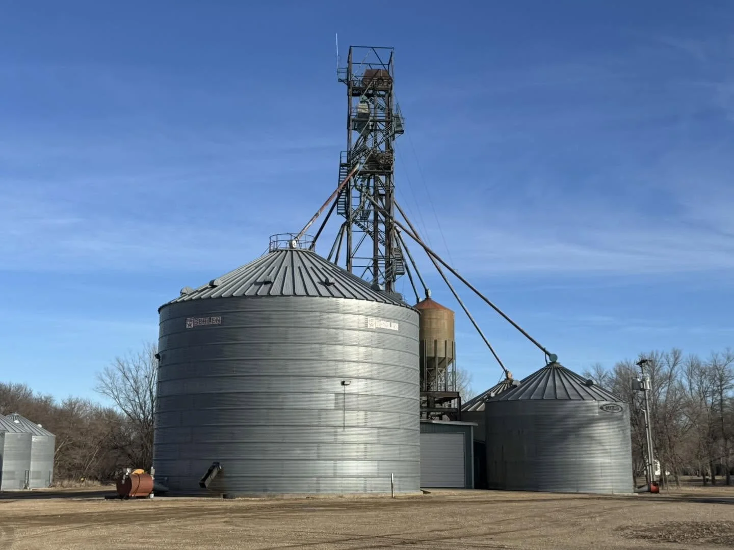 Two large metal grain silos with a tall metal grain elevator structure in the background and leafless trees in the distance under a blue sky.