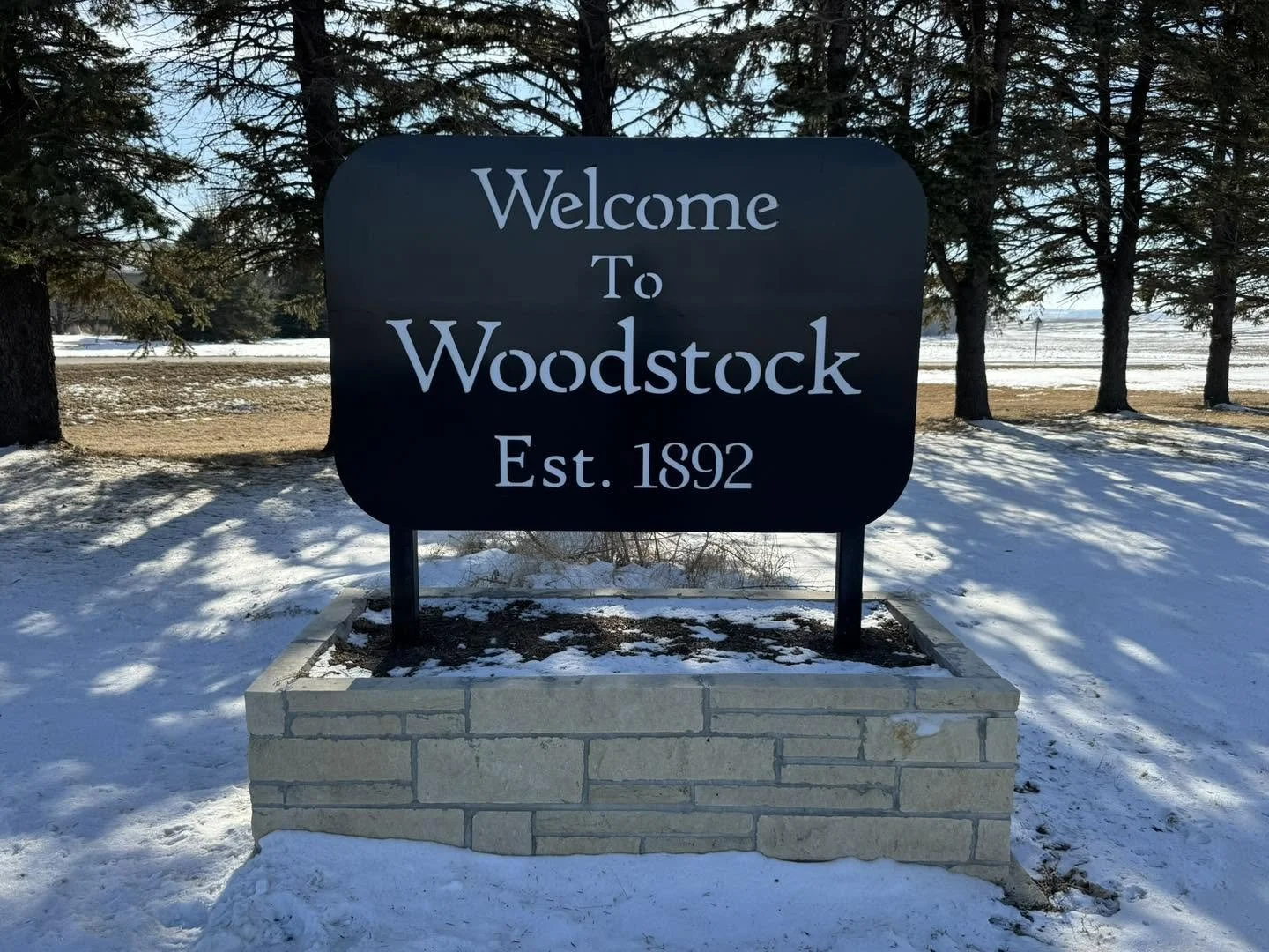 A black sign with white text that says, "Welcome to Woodstock, Est. 1892," mounted on two metal posts inside a stone planter, surrounded by snow and trees in the background.