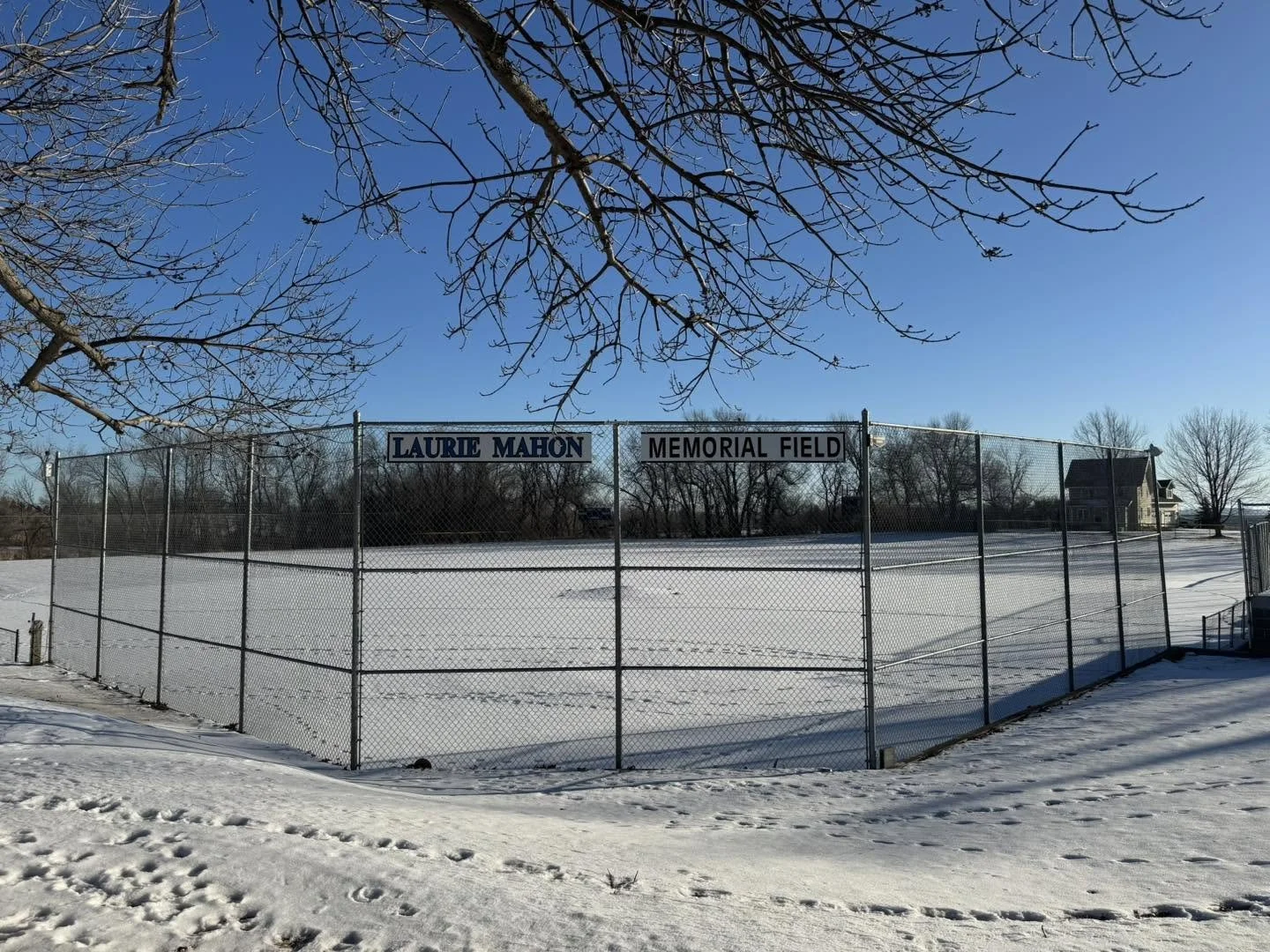 An outdoor ice hockey or skating rink enclosed by a chain-link fence, with a sign on the fence that reads 'LAURIE MAHON MEMORIAL FIELD.' The ground is covered with snow, and there are leafless trees and a house in the background under a clear blue sky.