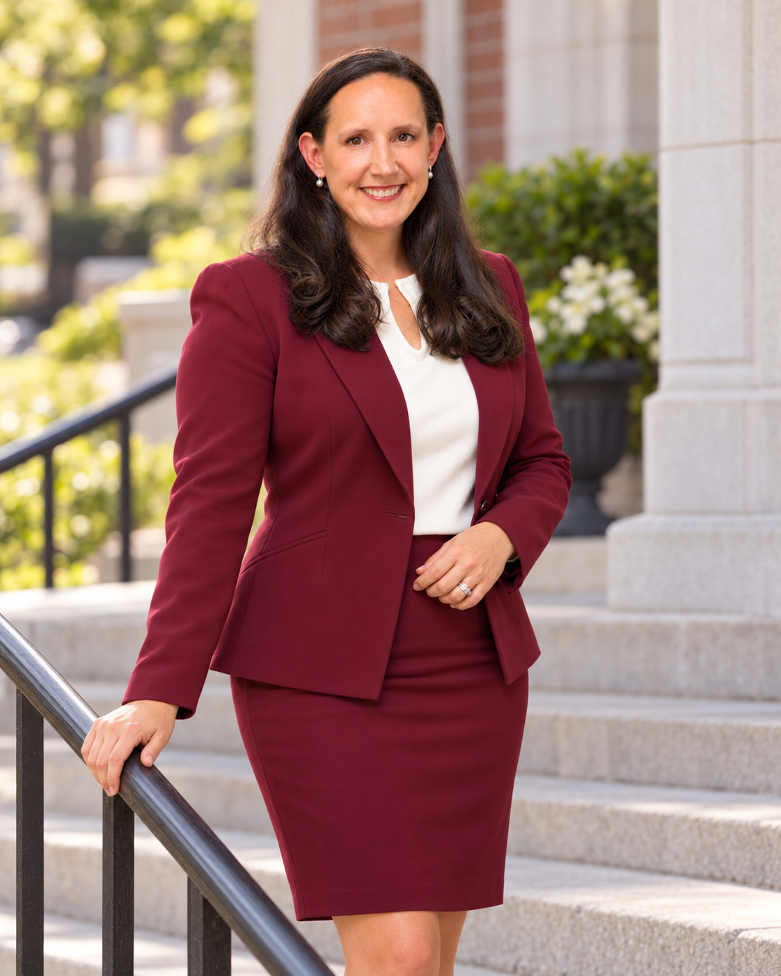 A woman with long brown hair and earrings wearing a white blouse and a maroon business suit, standing outdoors on steps near a building with greenery in the background.