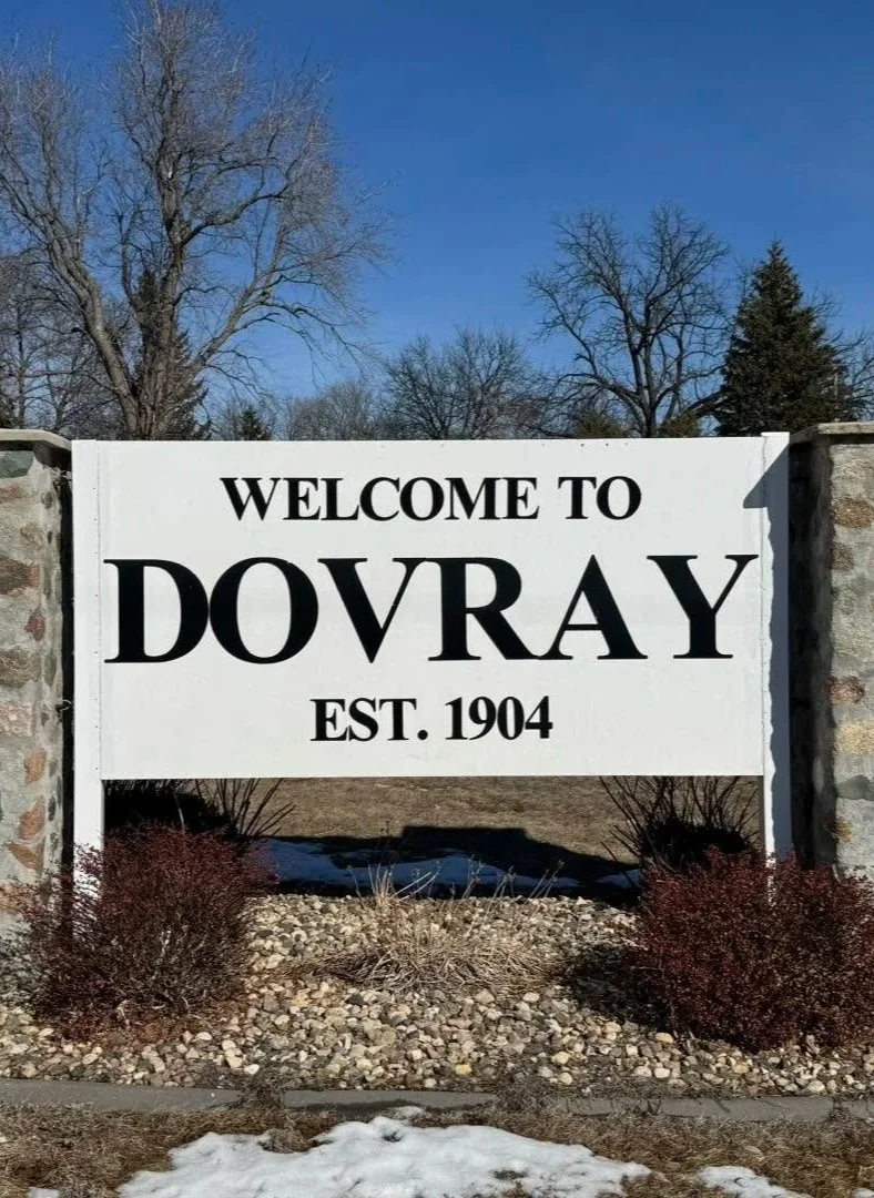 Welcome sign for Dovray, established in 1904, with stone pillars on each side, set in a landscaped area with shrubs and snow, under a clear blue sky.