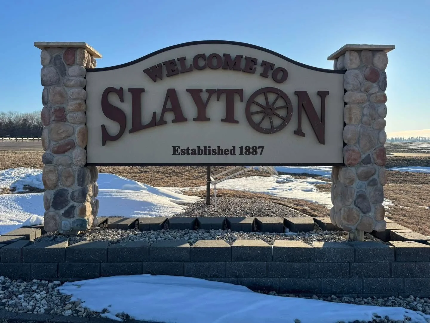 Stone sign welcoming visitors to Slayton, established in 1887, with a decorative wagon wheel in place of the letter 'O'.