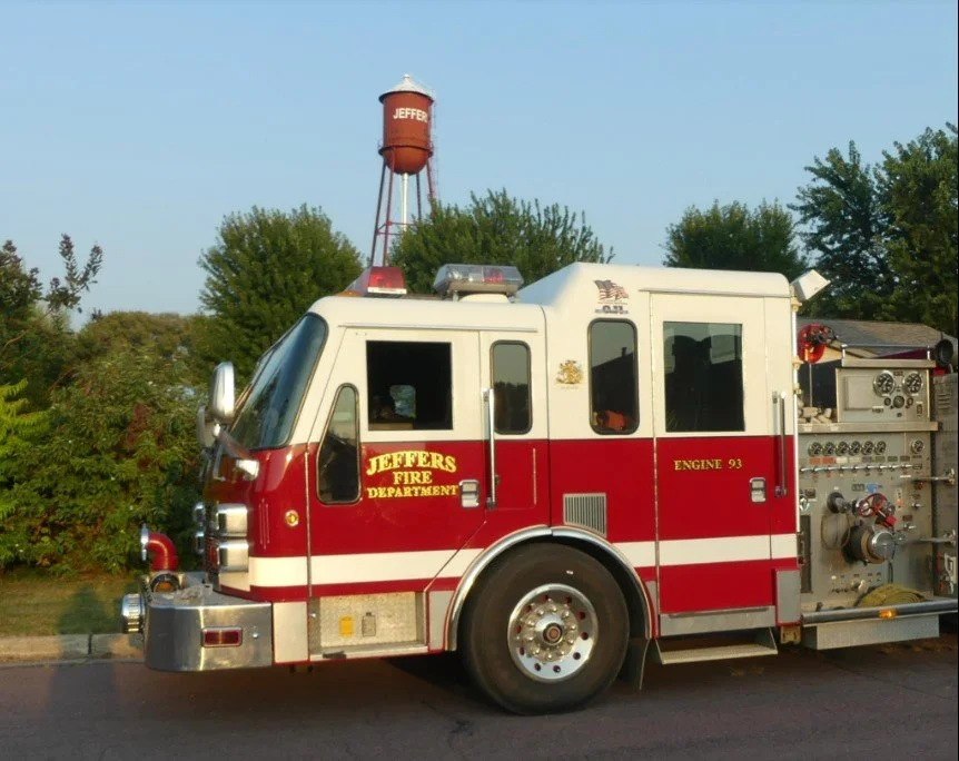 A red and white fire truck with the words 'Jeffers Fire Department' on the side, parked outdoors with trees and a water tower labeled 'Jeffers' in the background.