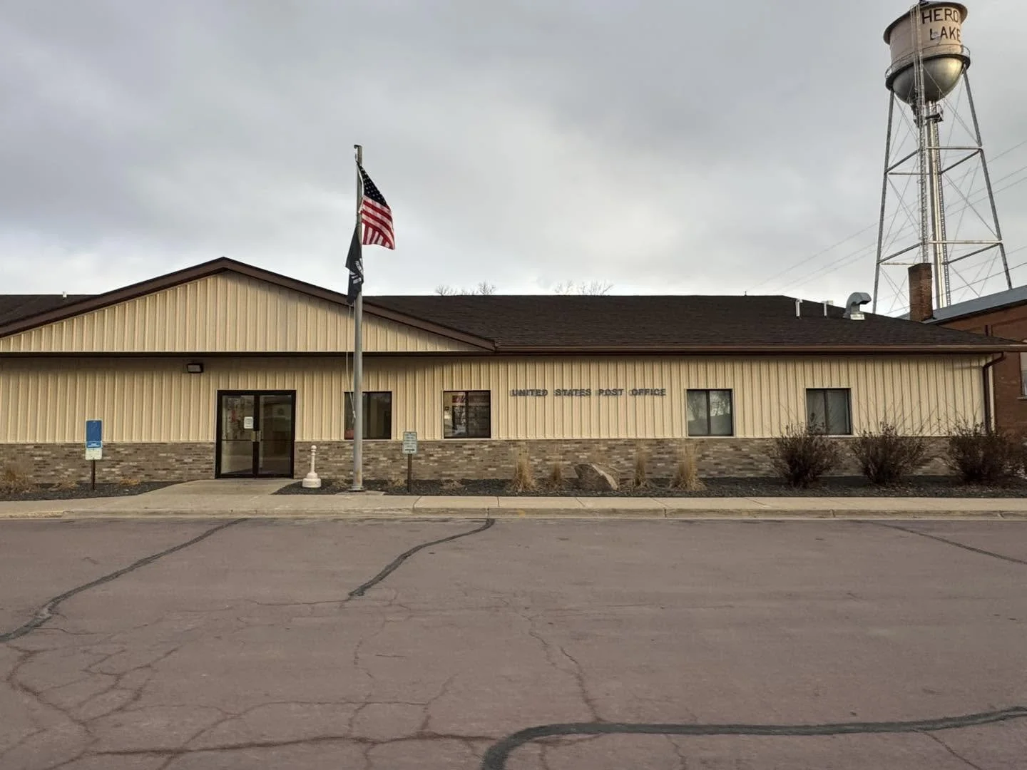 Small beige building with brick foundation labeled 'United States Post Office' with American flags outside, parking lot in front, and a water tower in the background.