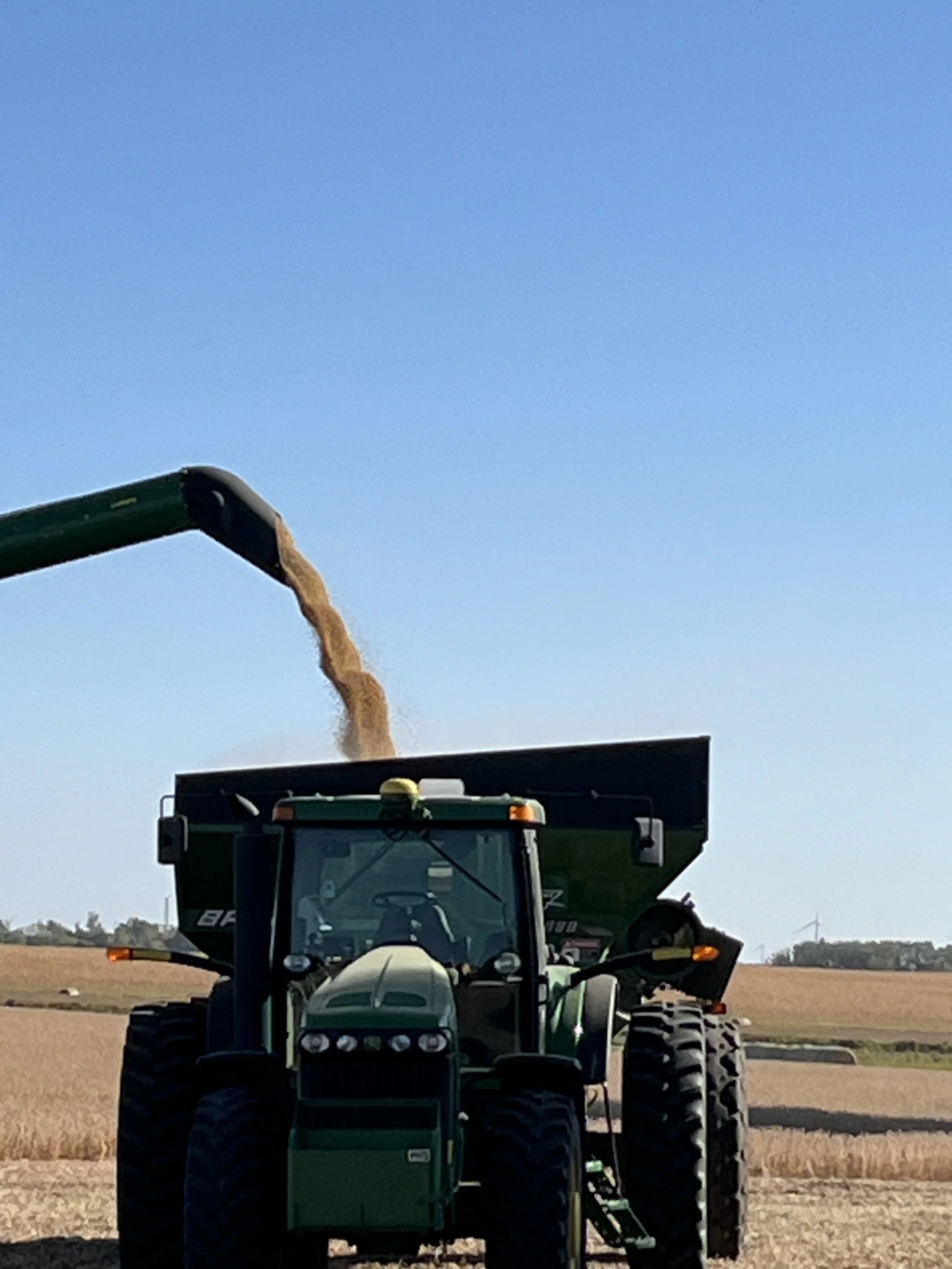 A tractor with a green and black body is in a field, with a large pipe extending from the tractor releasing a long stream of brownish material into the air against a blue sky background.
