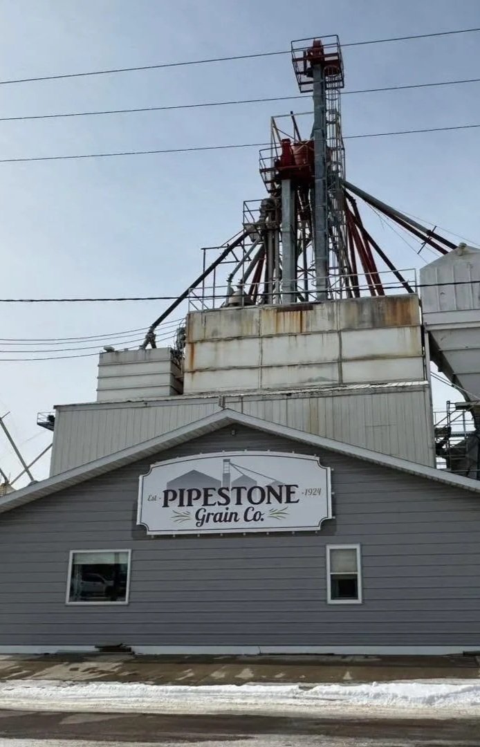 An industrial grain elevator building with a sign that reads "Est. Pipestone Grain Co. 1924" in front of a large metal structure with pipes and towers.