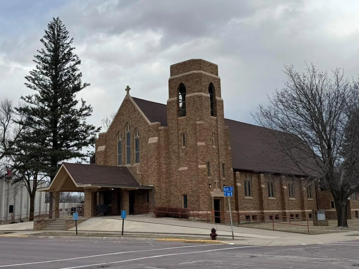 A brick church with a tall bell tower, pointed roof, stained glass windows, surrounded by leafless trees on a cloudy day, with street signs and parking in the foreground.